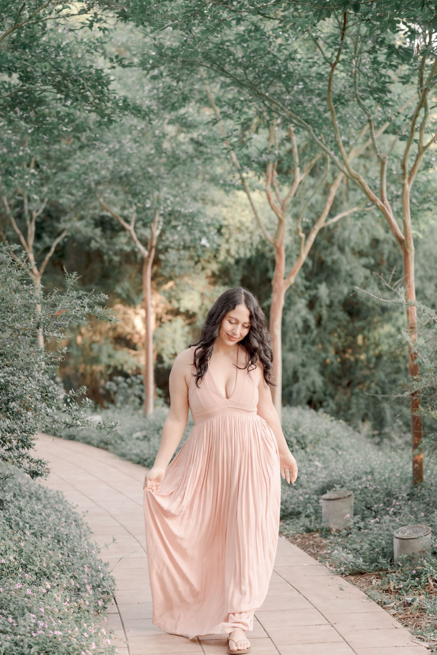 A woman in a flowing blush pink maxi dress walks along a tree-lined garden pathway.