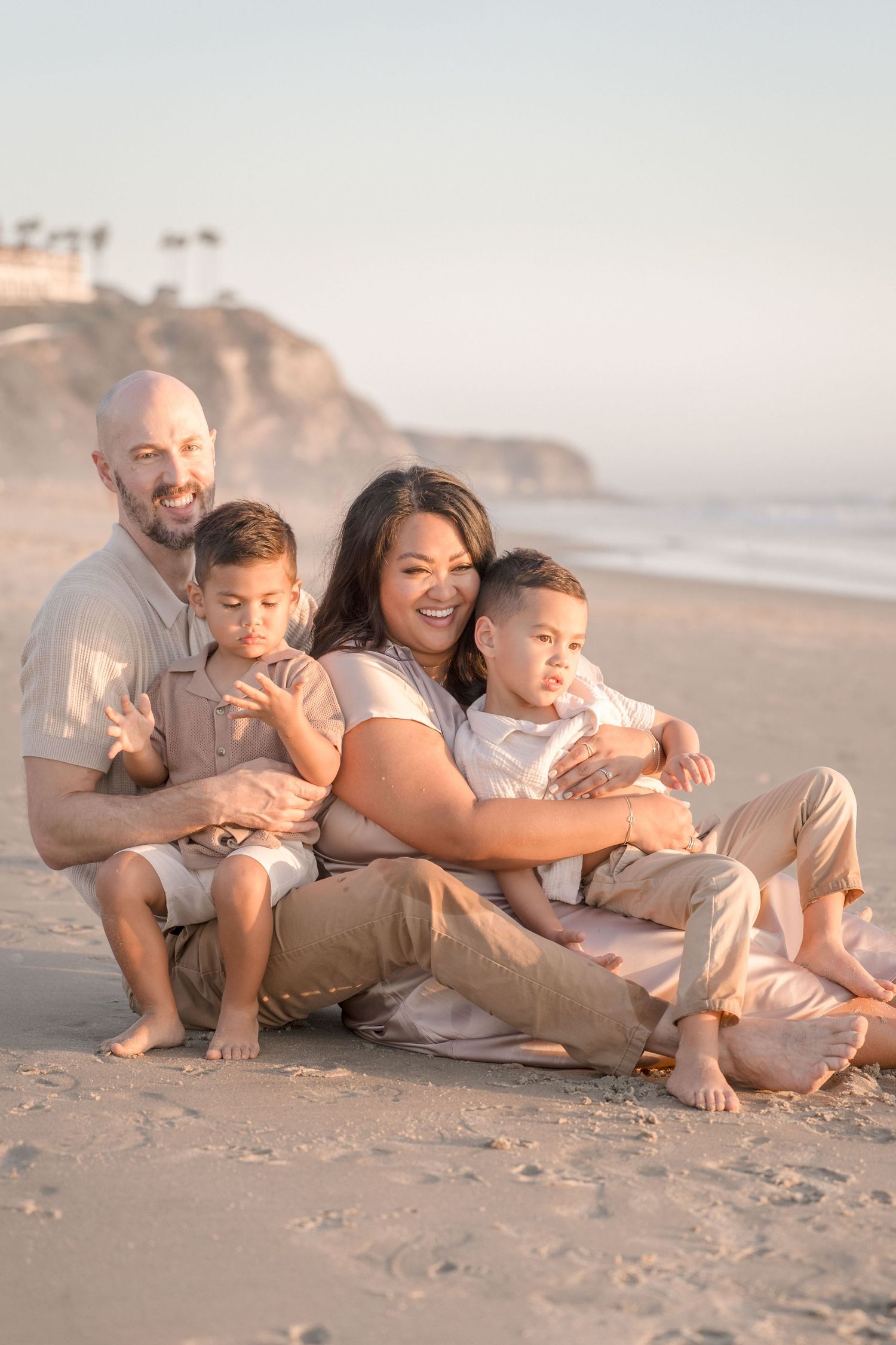 Happy family of four sitting together on a sandy beach, laughing and cuddling at golden hour.