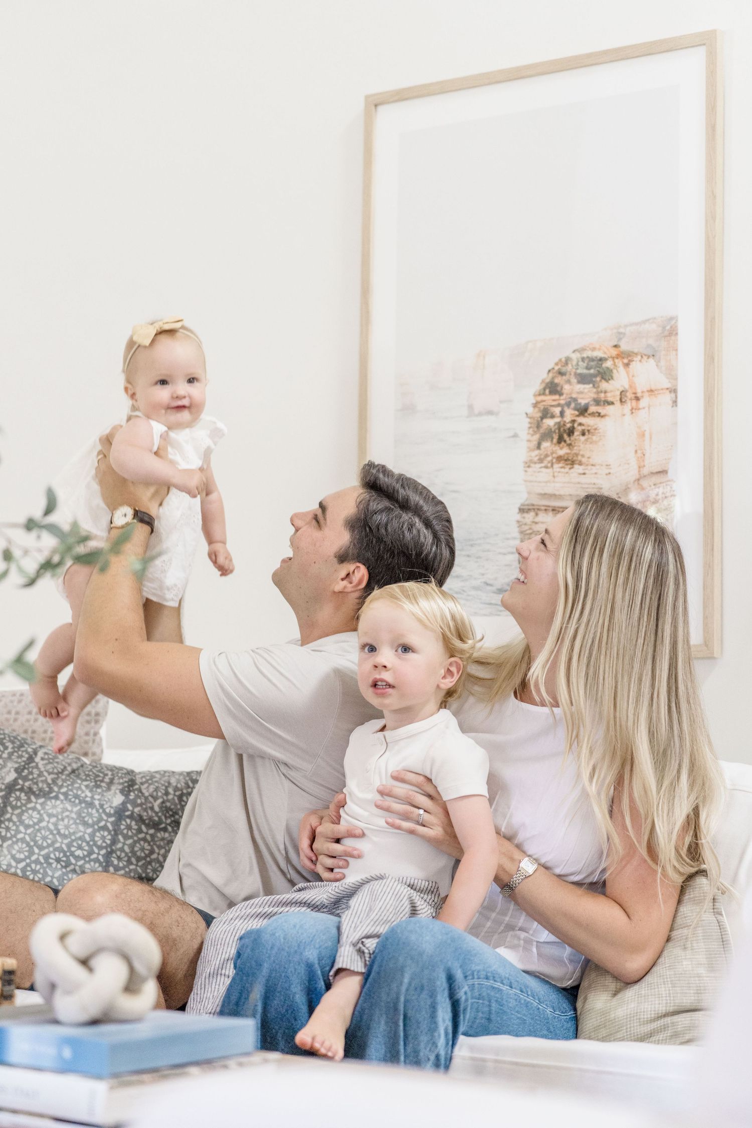Joyful family of four on a couch, father raising smiling baby girl while mother holds toddler son.
