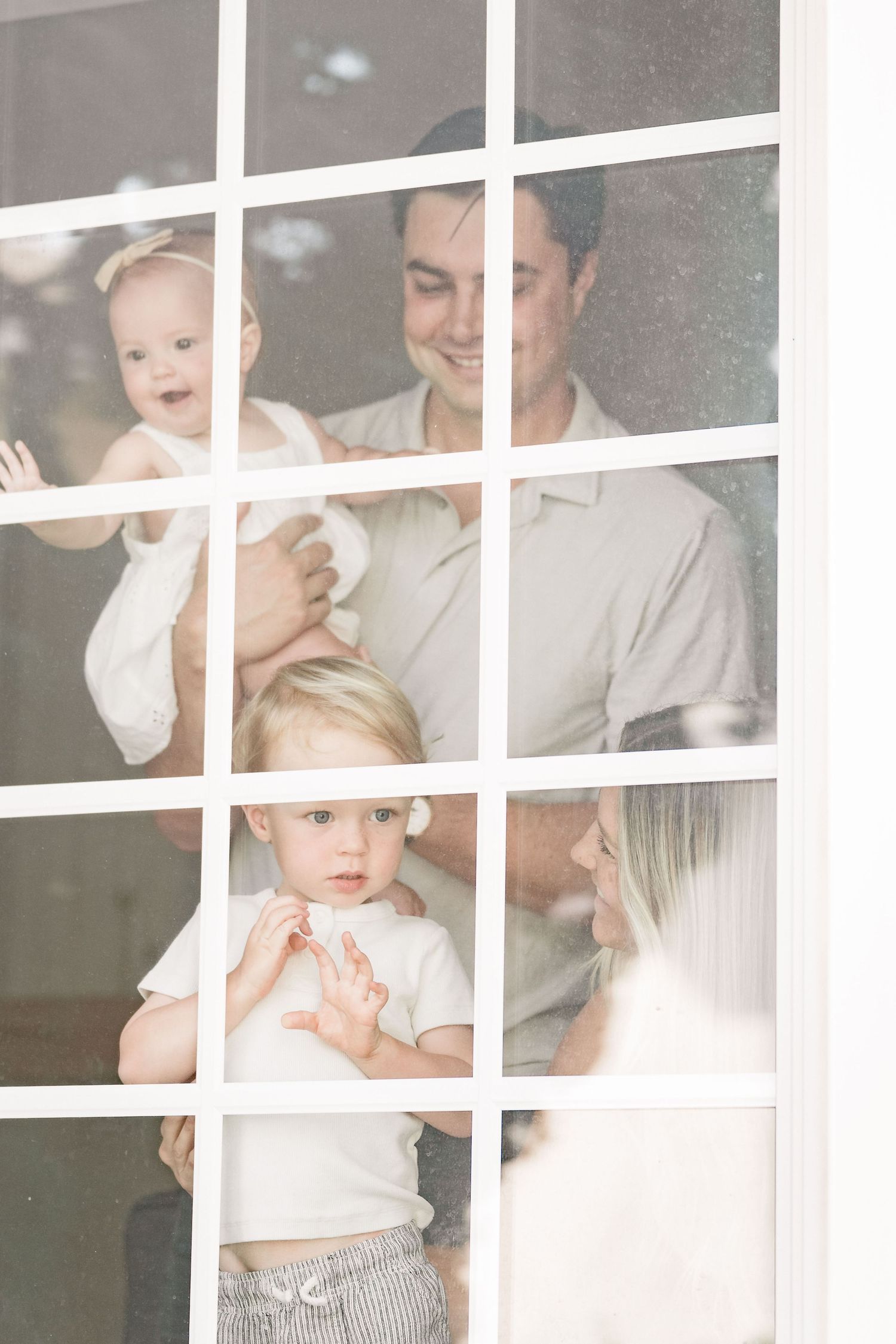 A happy family with two young children is seen smiling through a white-framed window.