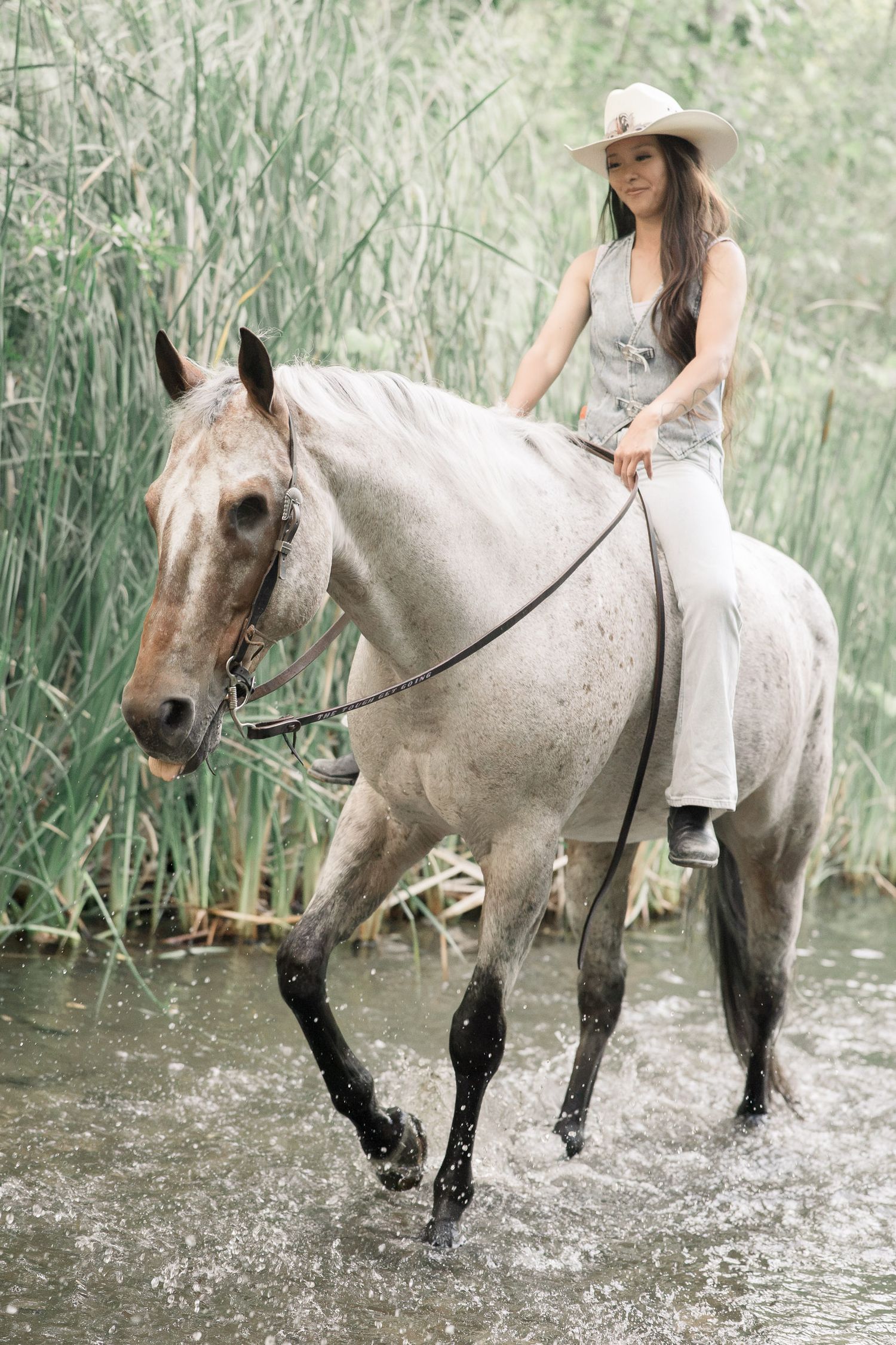 Woman in cowboy hat riding a grey horse through a shallow river surrounded by tall green reeds.
