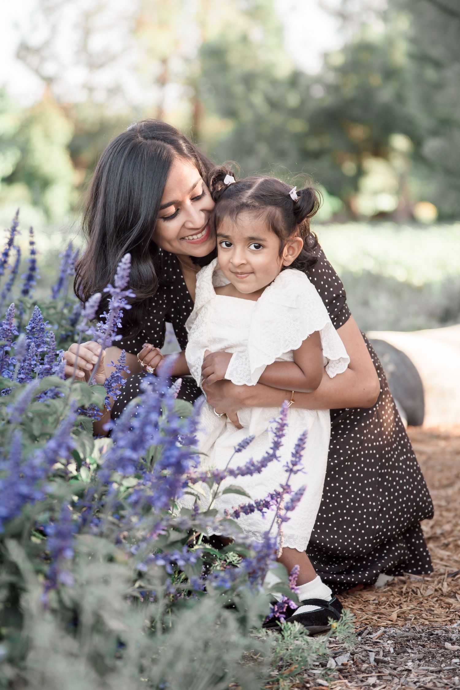 A smiling mother in a polka dot dress hugs her young daughter near purple lavender flowers in a lush green park.