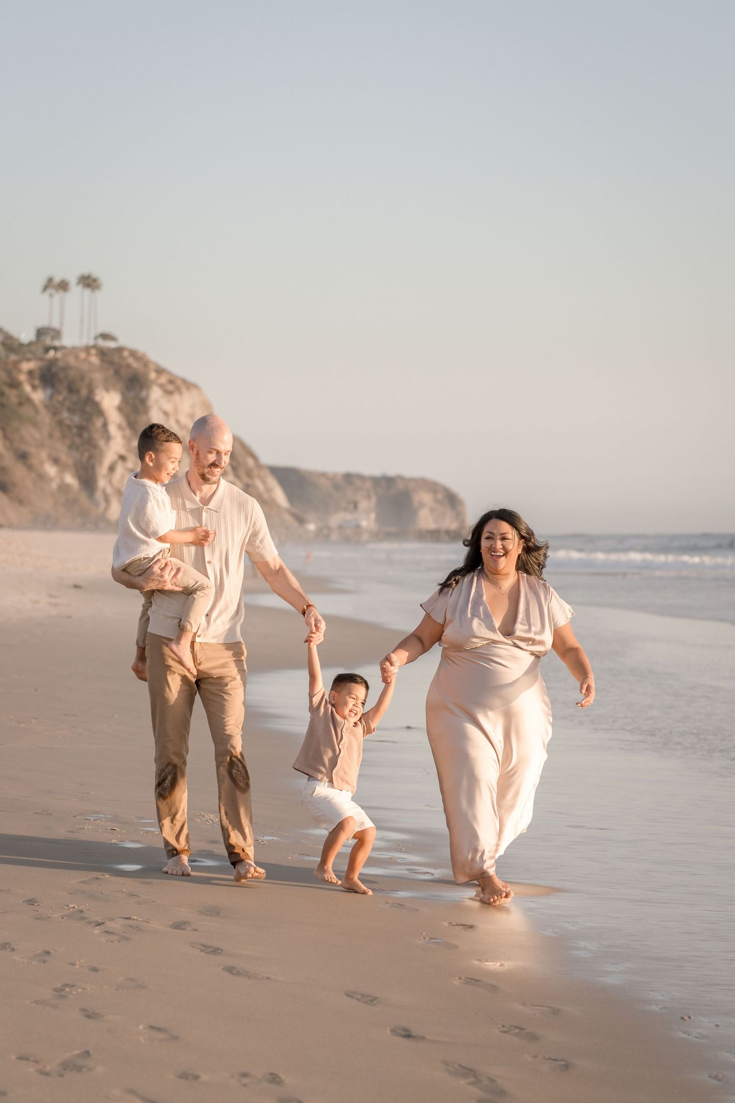 Happy family of four walking along a sunny beach, parents swinging young child between them.