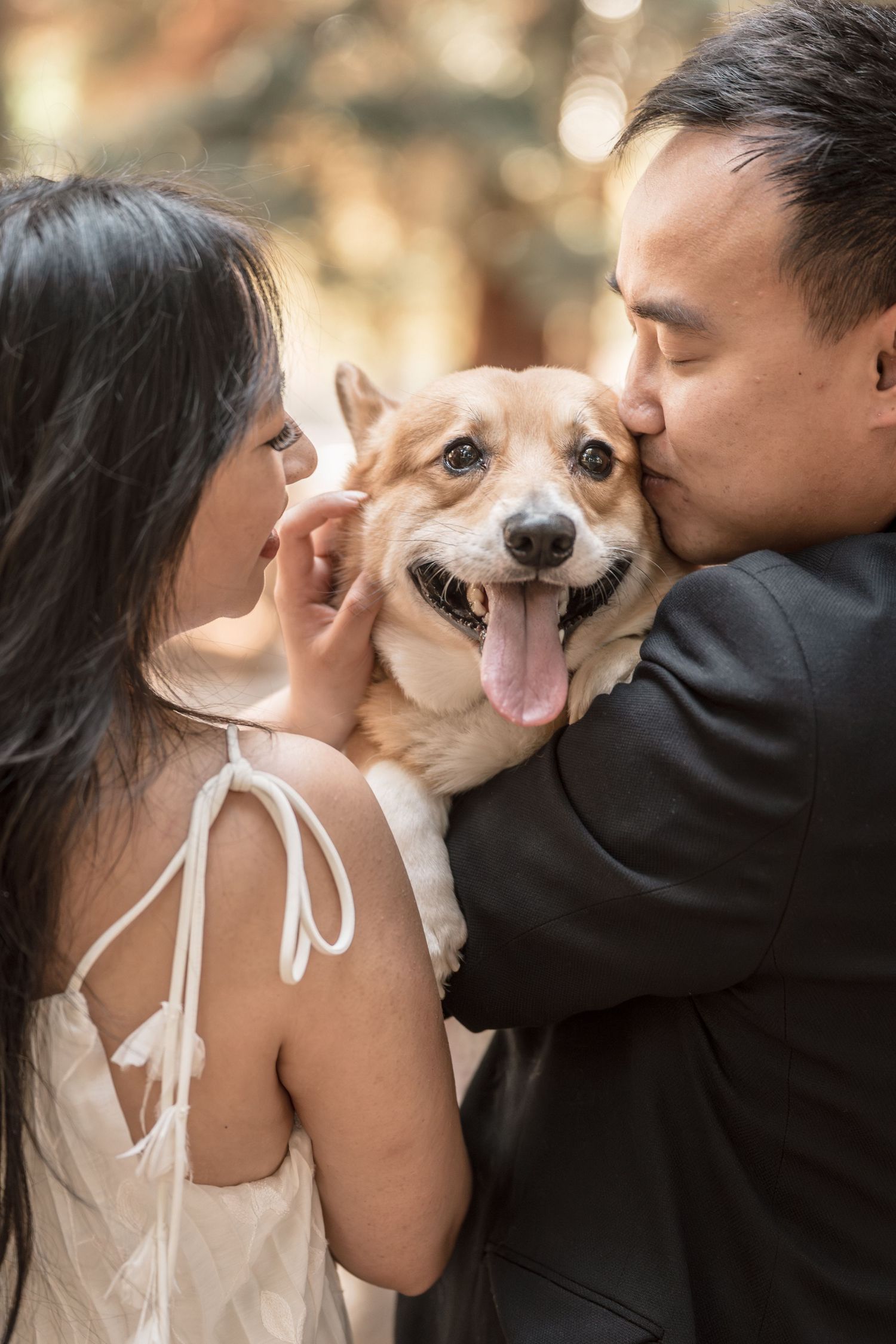 An Asian couple lovingly kisses their happy Corgi dog on both sides of its face outdoors.
