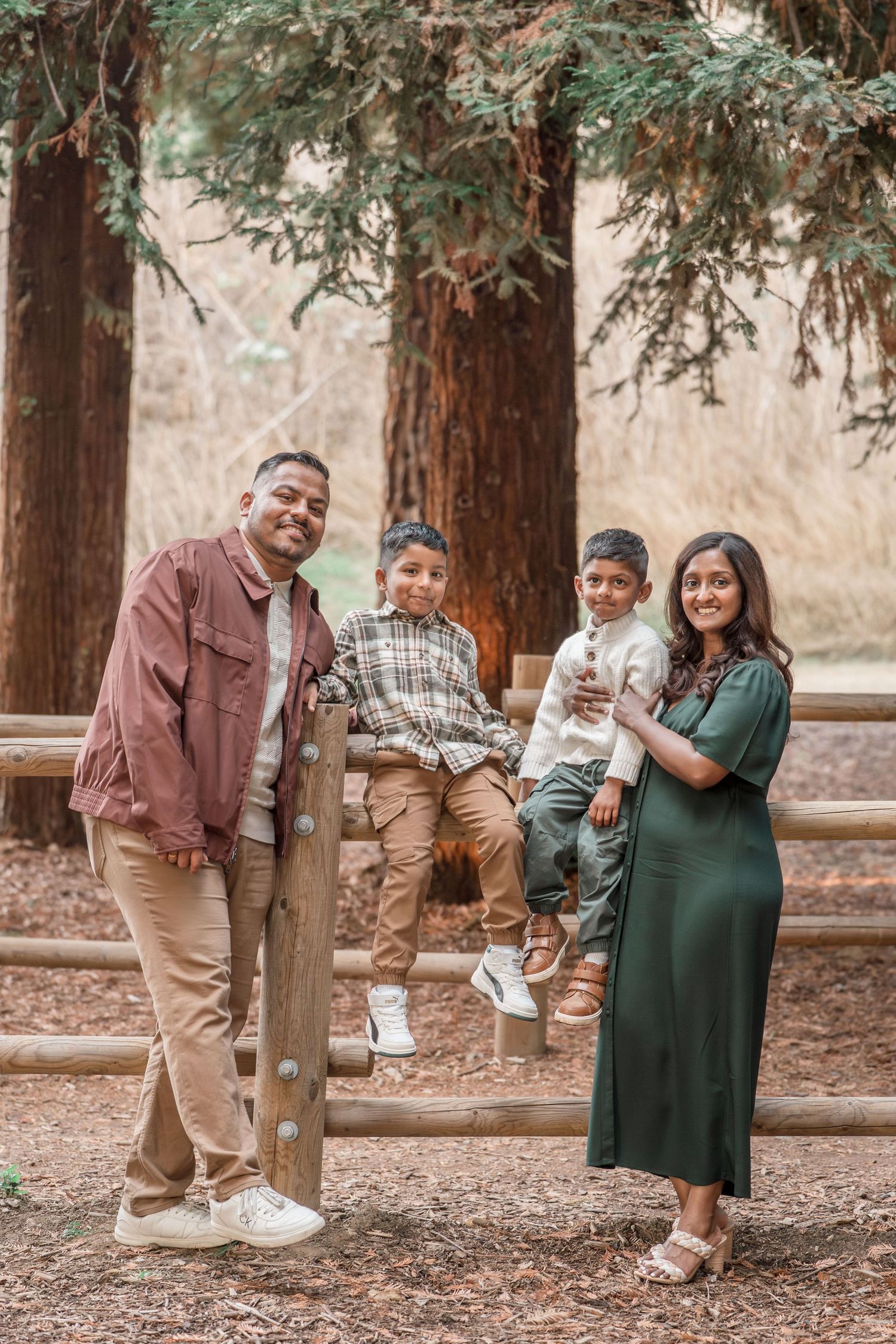 A South Asian family of four smiles together by a wooden fence surrounded by tall redwood trees.