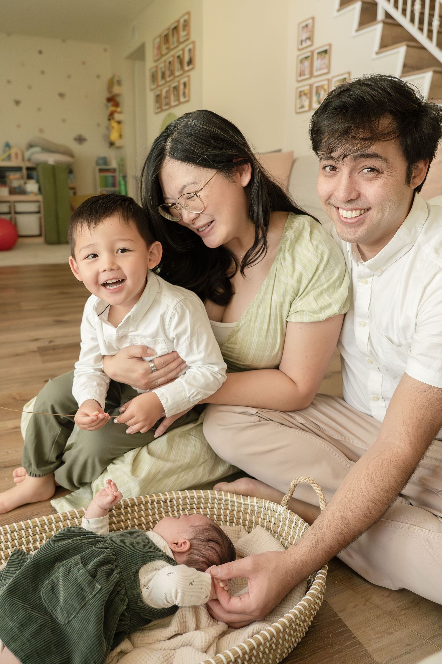 Happy family of four sitting on floor, parents smiling with toddler and newborn baby in wicker basket at home.