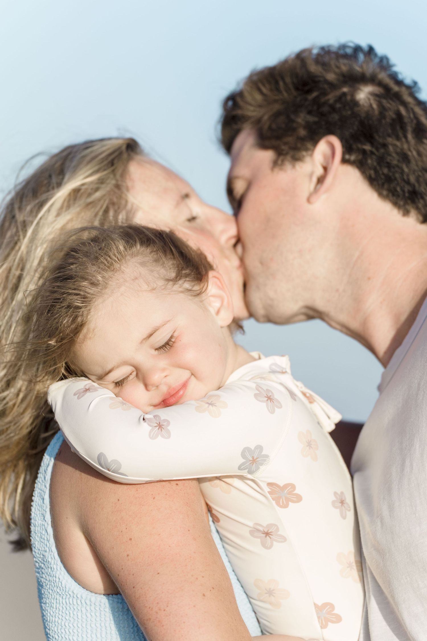 Two parents lovingly kiss their smiling young daughter on the cheeks outdoors against a blue sky.