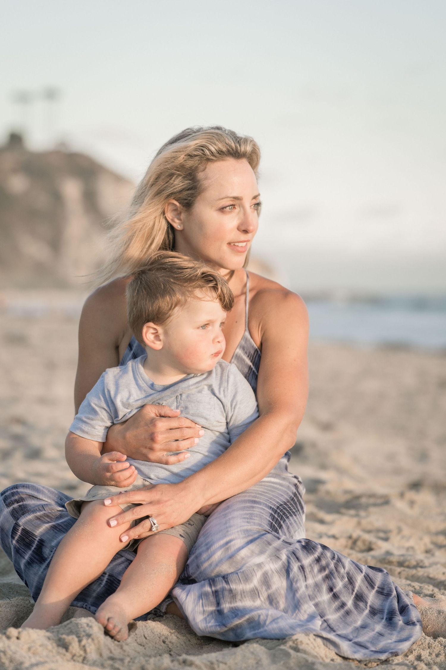 A blonde mother in a plaid dress holds her young son while sitting on a sandy beach at sunset.