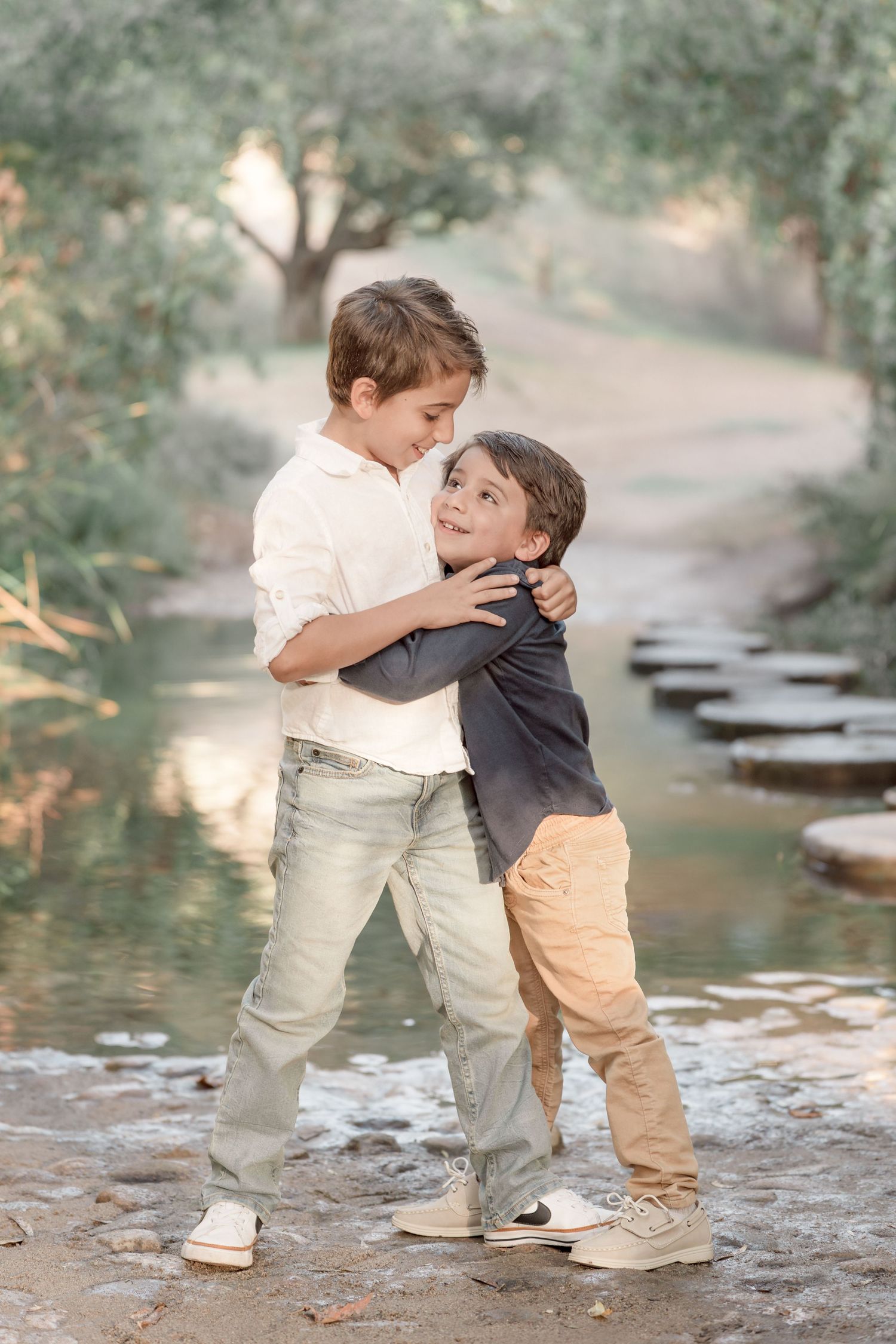 Two young brothers share a warm hug while standing near a peaceful tree-lined creek in soft natural light.
