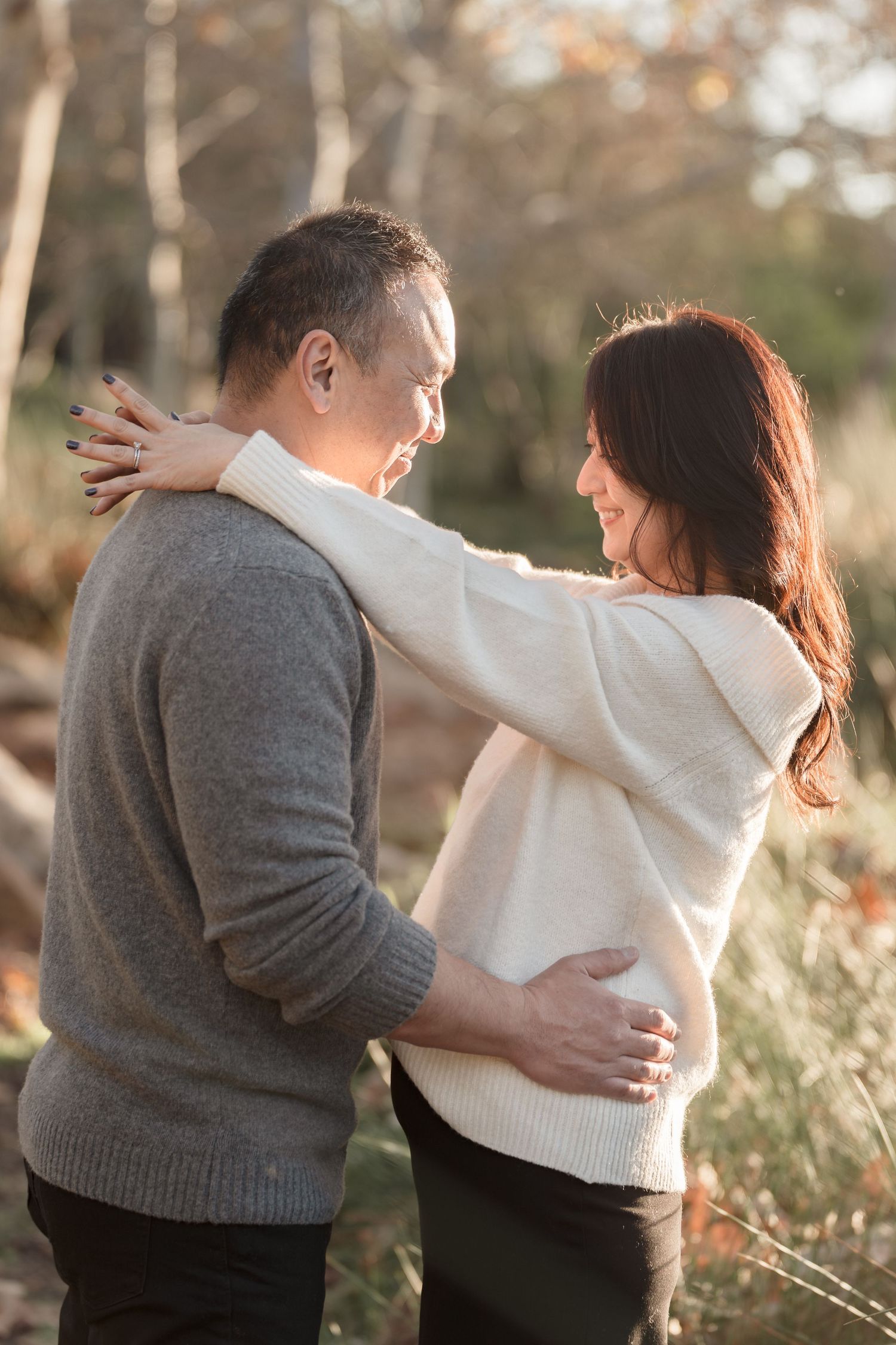 A couple embraces outdoors in autumn, the woman resting her arm on the man's shoulder while he holds her waist.