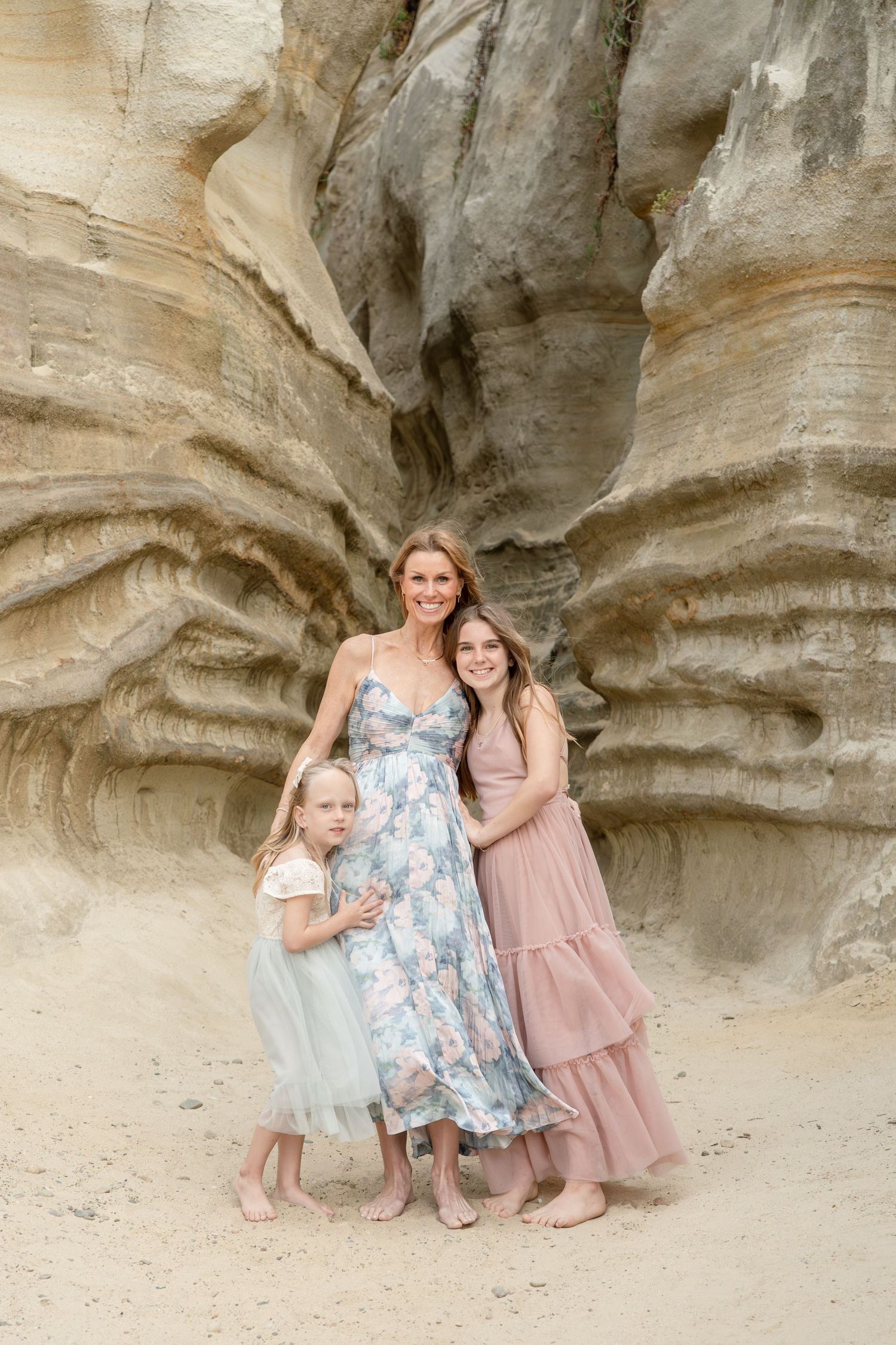 A woman and two girls in pastel dresses pose barefoot before layered sandstone rock formations.