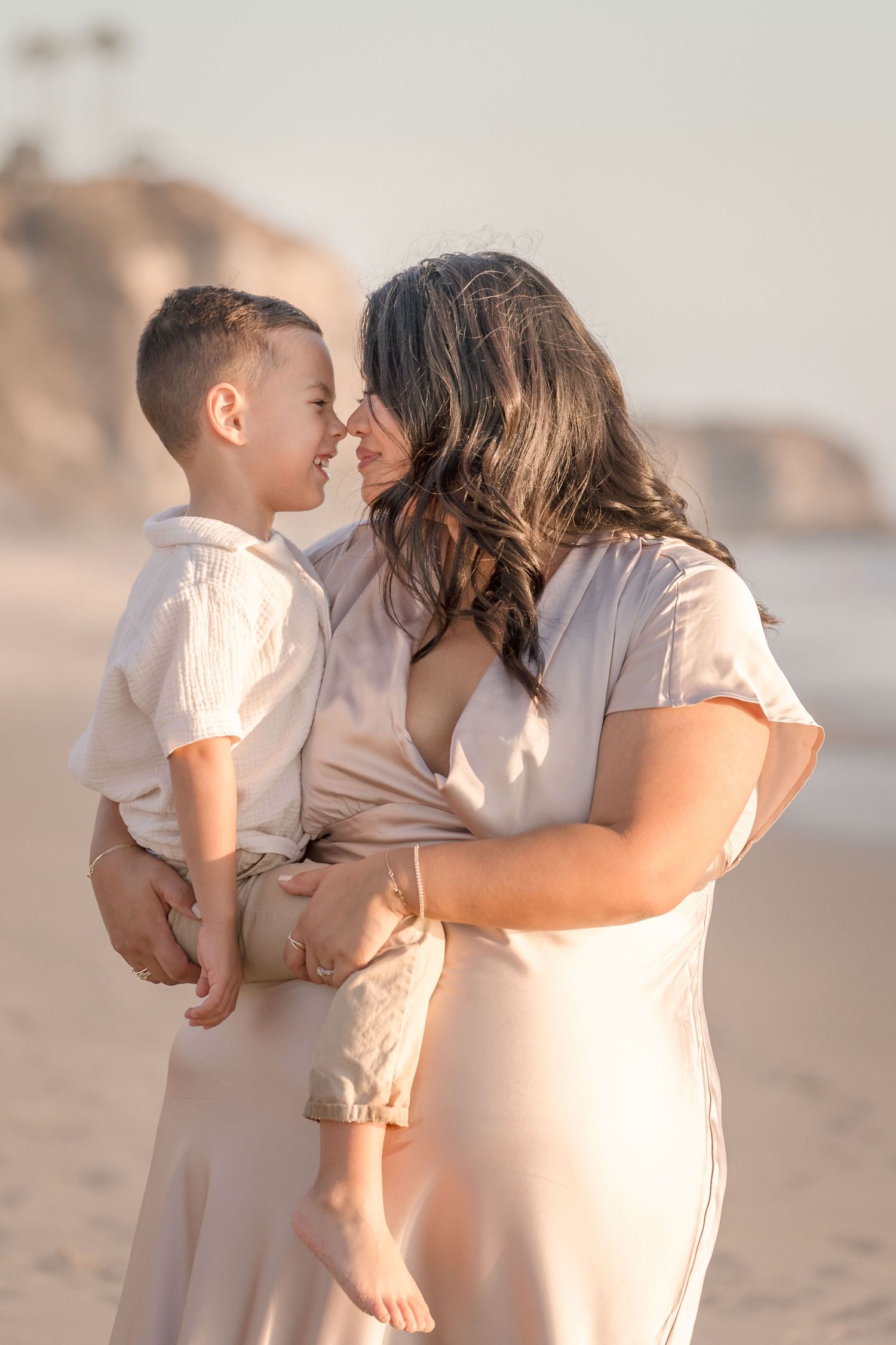 A smiling mother in a blush dress holds her young son as they touch foreheads on a beach.