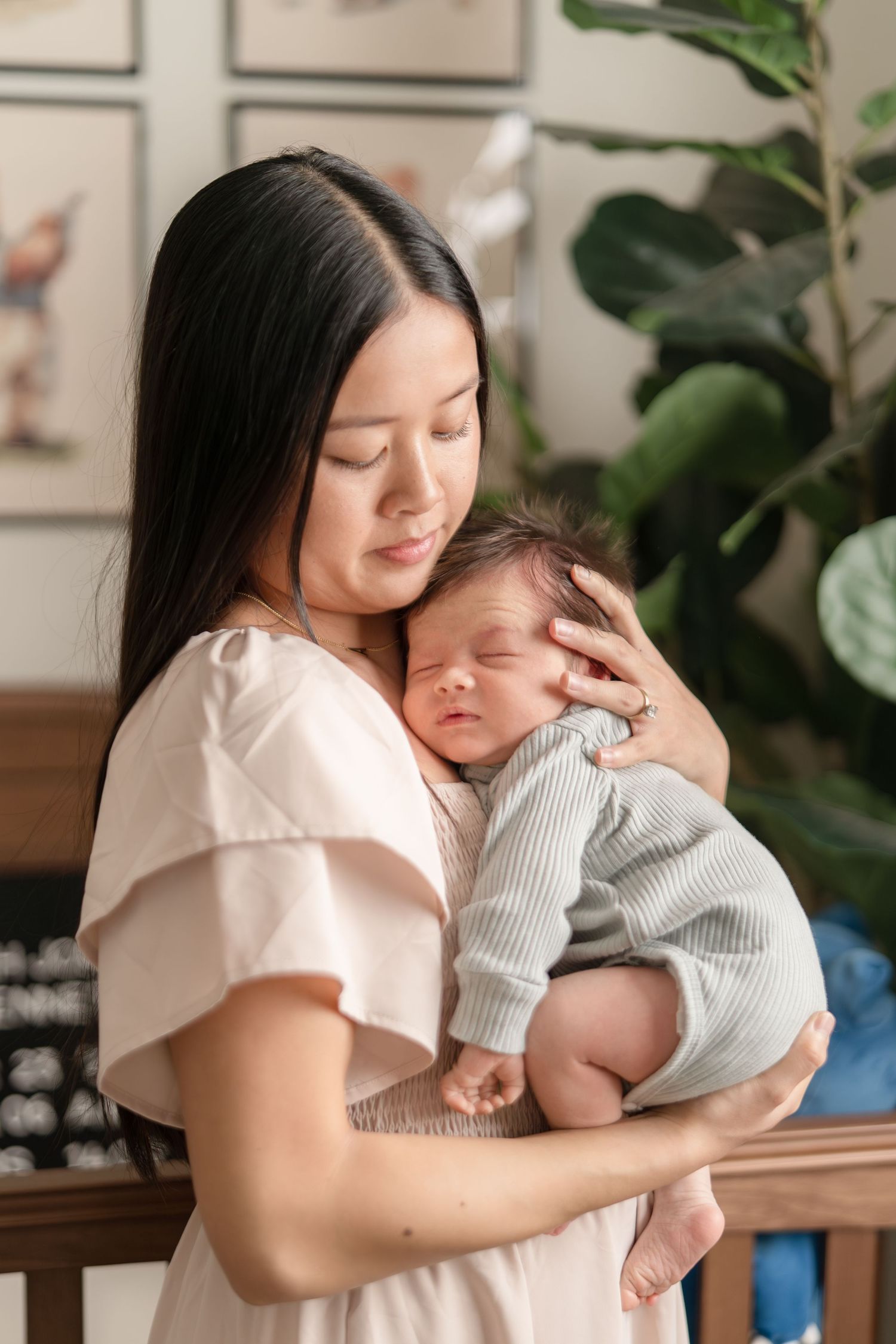 Asian mother in pink dress tenderly holds sleeping newborn baby in a cozy nursery.