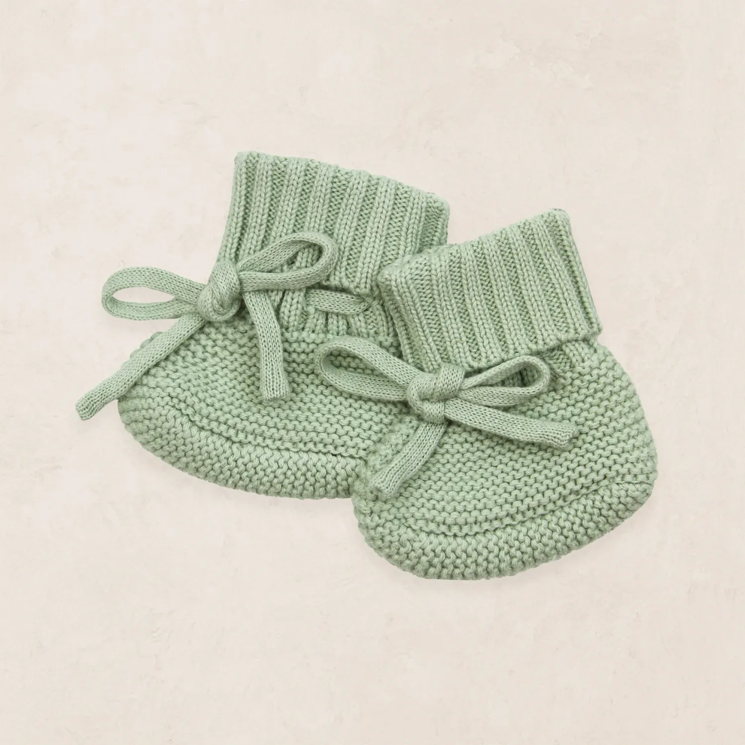 Sage green knit baby booties with bow ties displayed on a soft neutral background.