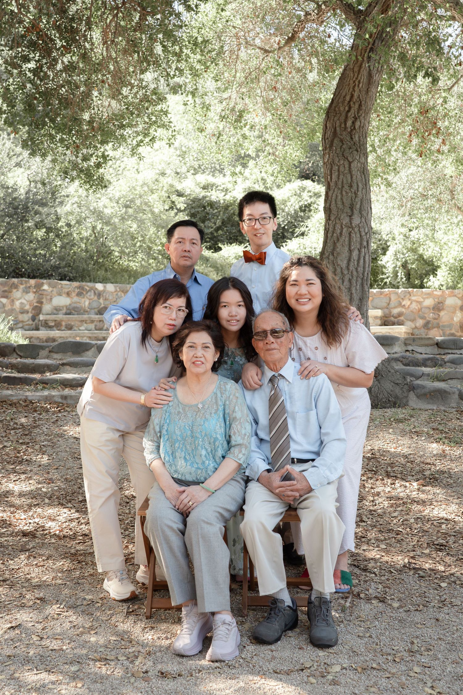 Multi-generational Asian family posing together outdoors under a large tree in a park setting.