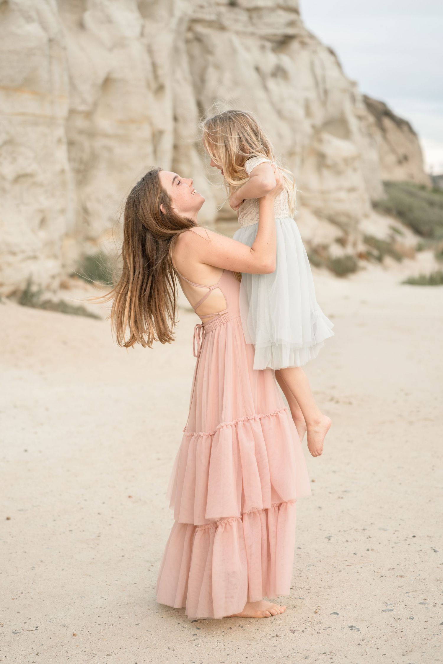 A mother in a pink tiered dress lifts her young daughter in a white dress on a sandy beach near rocky cliffs.