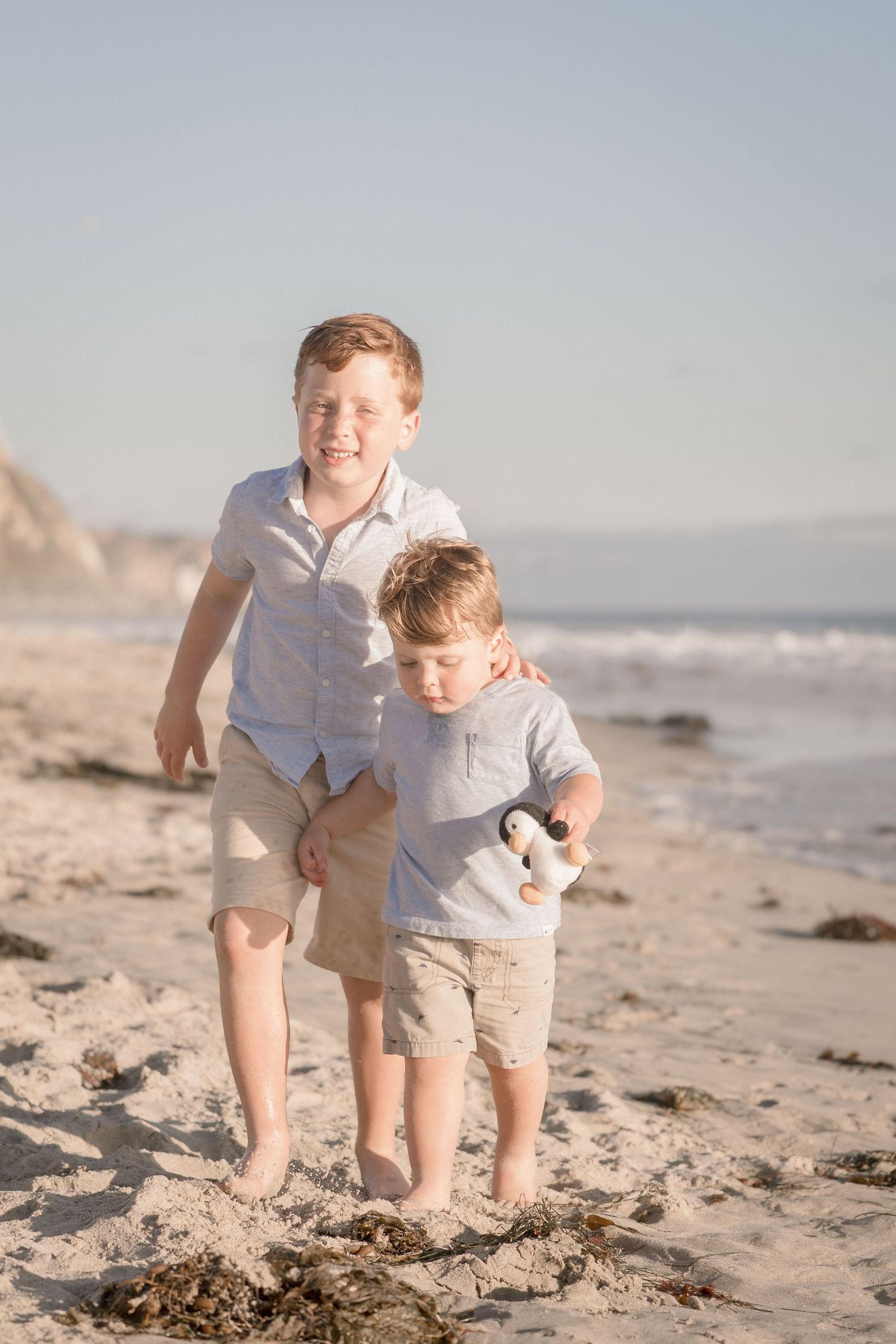 Two young red-haired brothers smile and pose together on a sunny beach near the ocean.