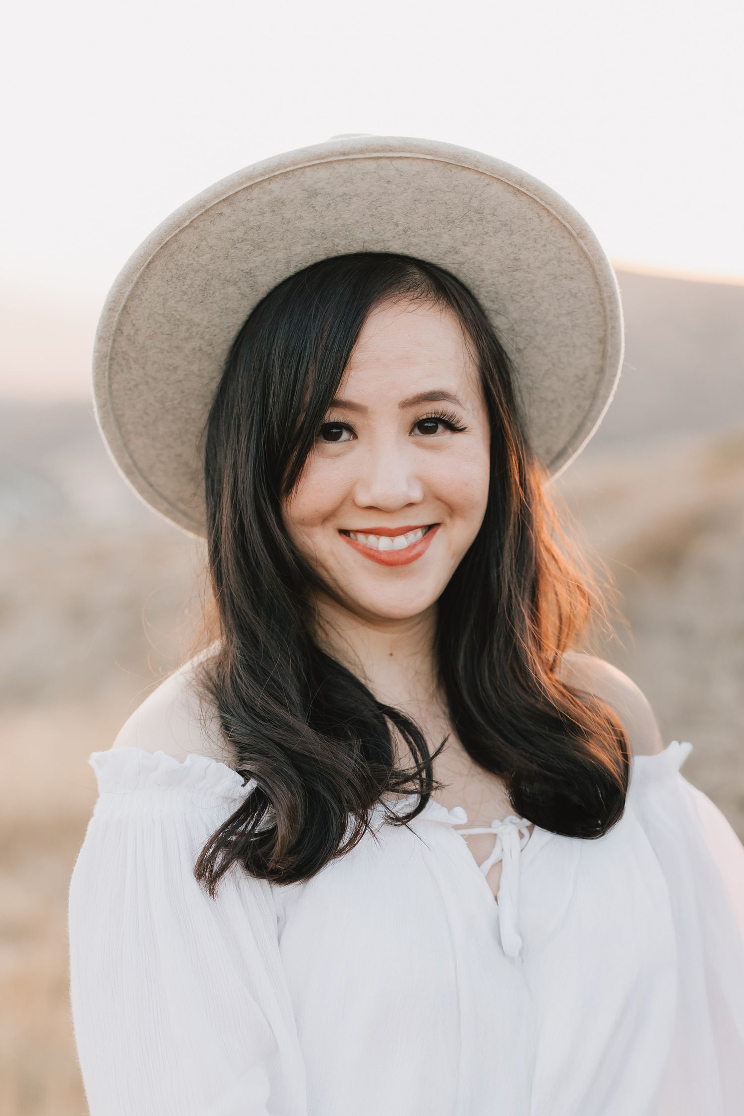 A smiling young woman wearing a wide-brim beige hat and white off-shoulder top in a sunny outdoor setting.