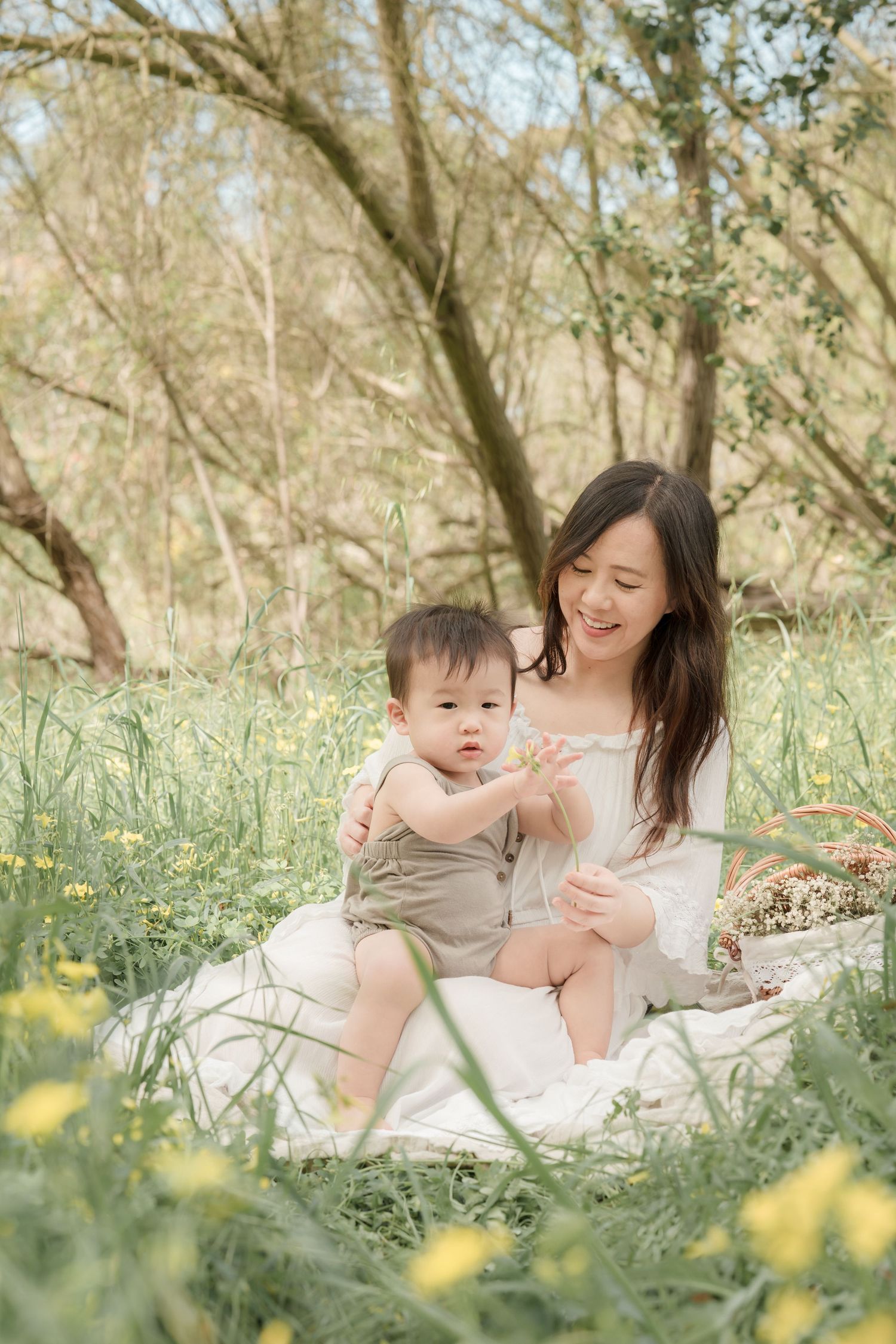 A smiling Asian mother sits on a blanket in a meadow with yellow wildflowers, holding her baby in a green romper.