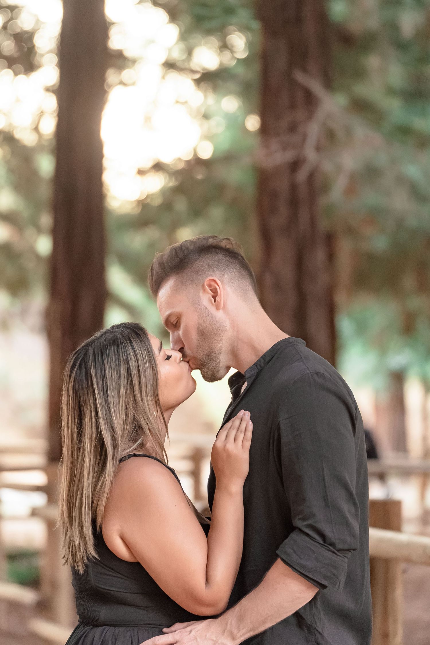 Couple sharing a romantic kiss surrounded by tall redwood trees in warm golden sunlight.