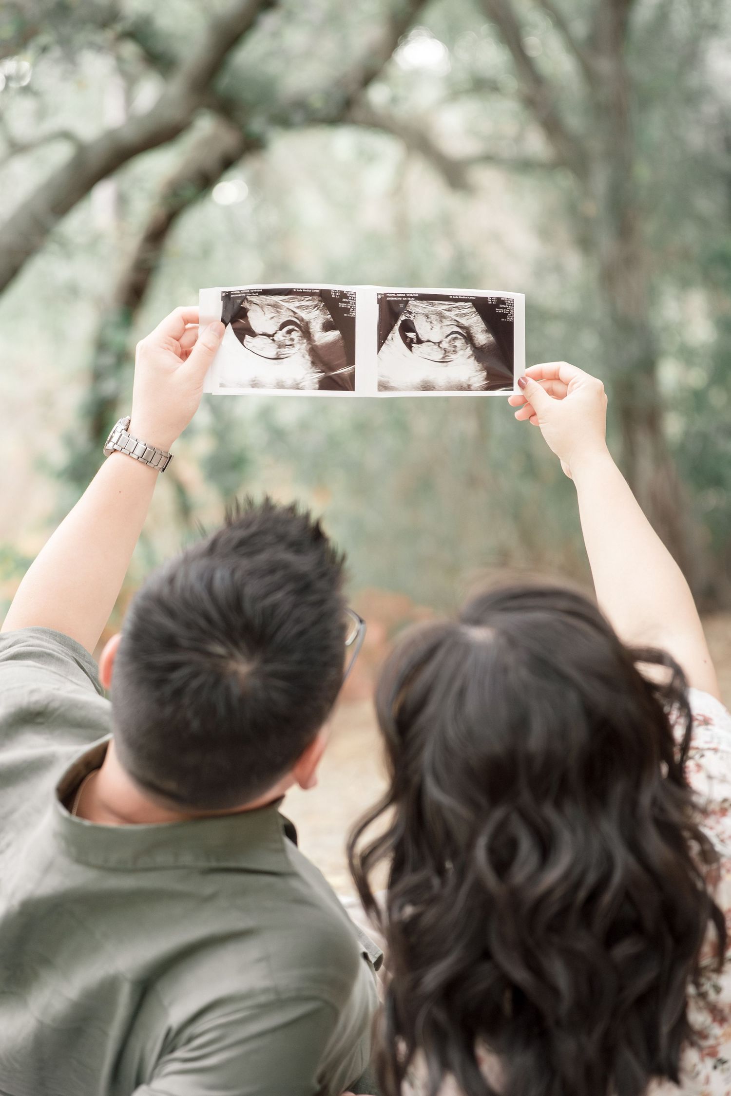 Couple holding up two ultrasound photos together outdoors under oak trees, viewed from behind.