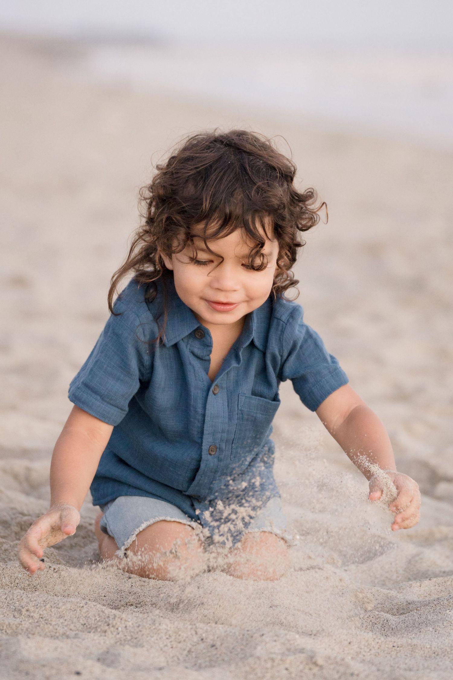 A curly-haired toddler boy in a blue shirt happily plays in the sand at the beach.