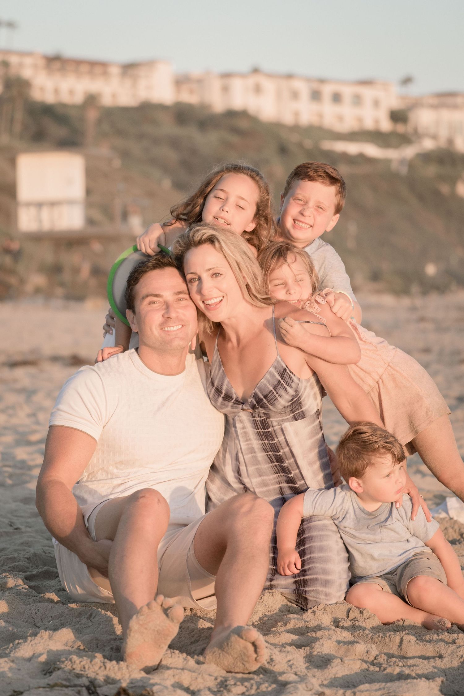 Happy family of six posing together on a sandy beach at sunset with coastal buildings behind them.