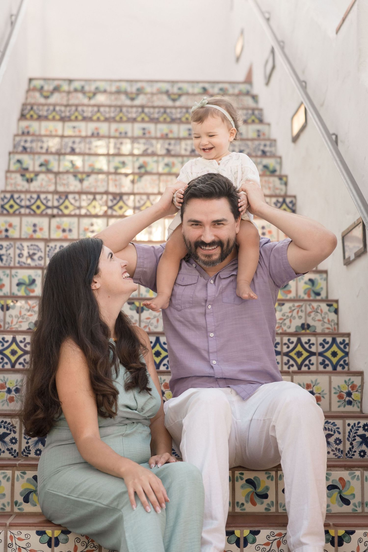 Happy family with toddler on dad's shoulders sitting on colorful tiled staircase.