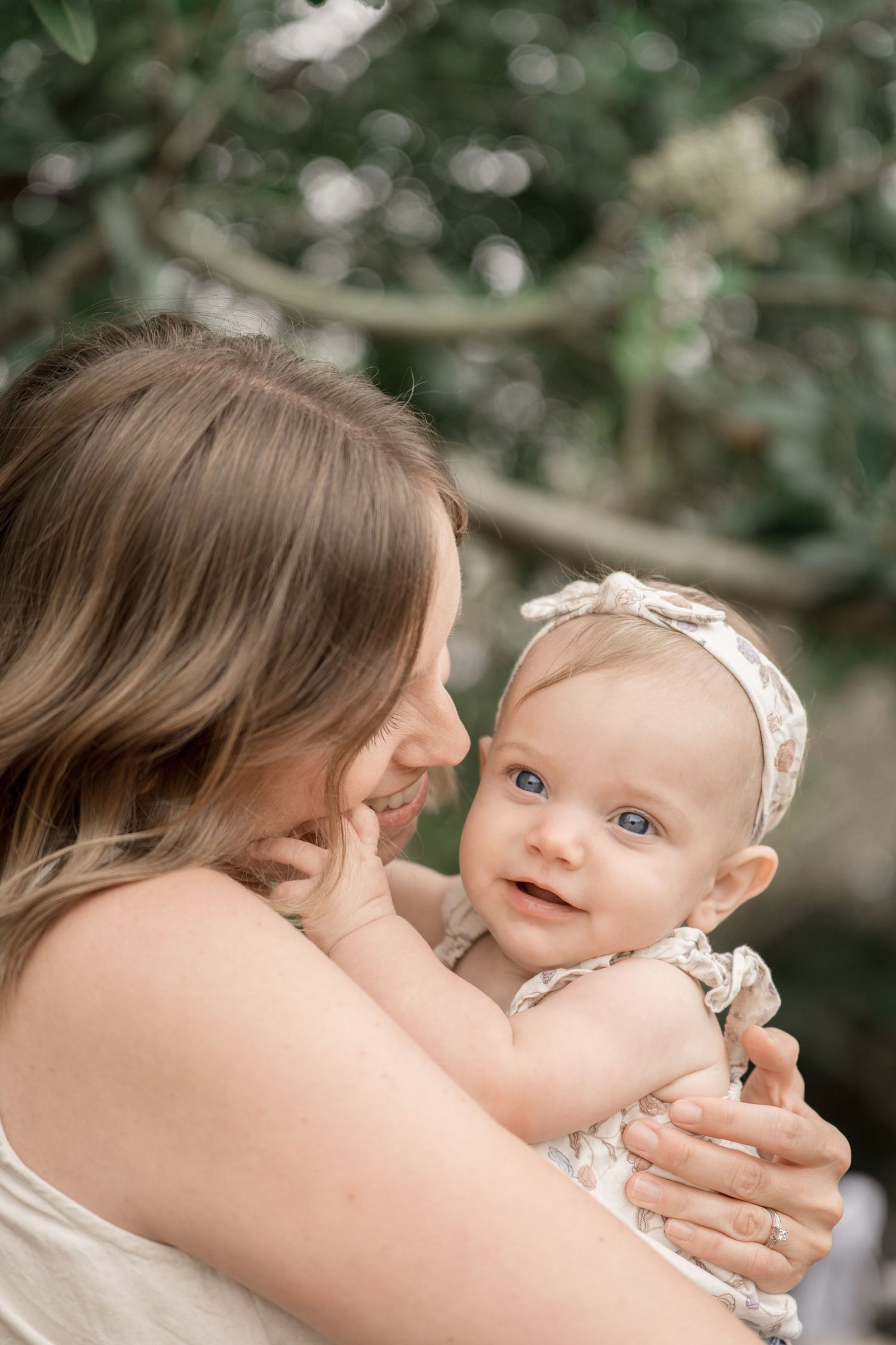 A mother tenderly holds her smiling blue-eyed baby girl wearing a floral outfit and white headband outdoors.
