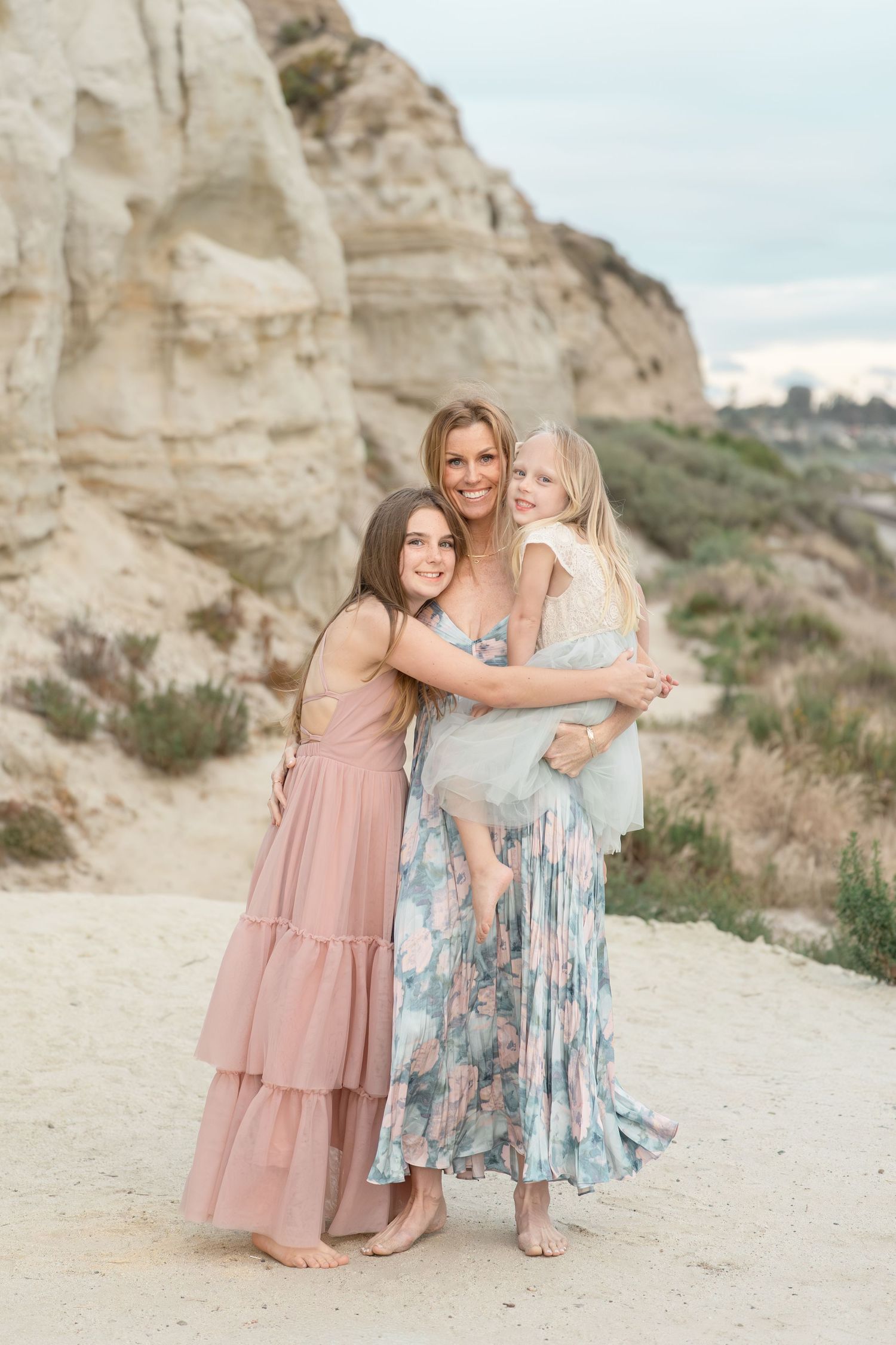 A mother and two daughters in pastel dresses embrace outdoors near sandy cliffs in a warm family portrait.