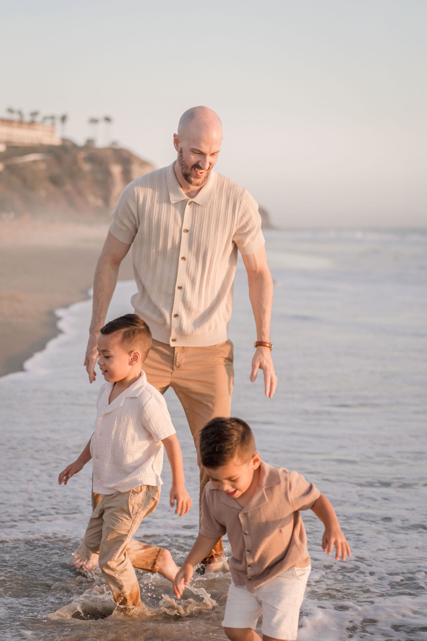 Bald father laughing as two young boys splash in ocean waves at sunset on a sandy beach.