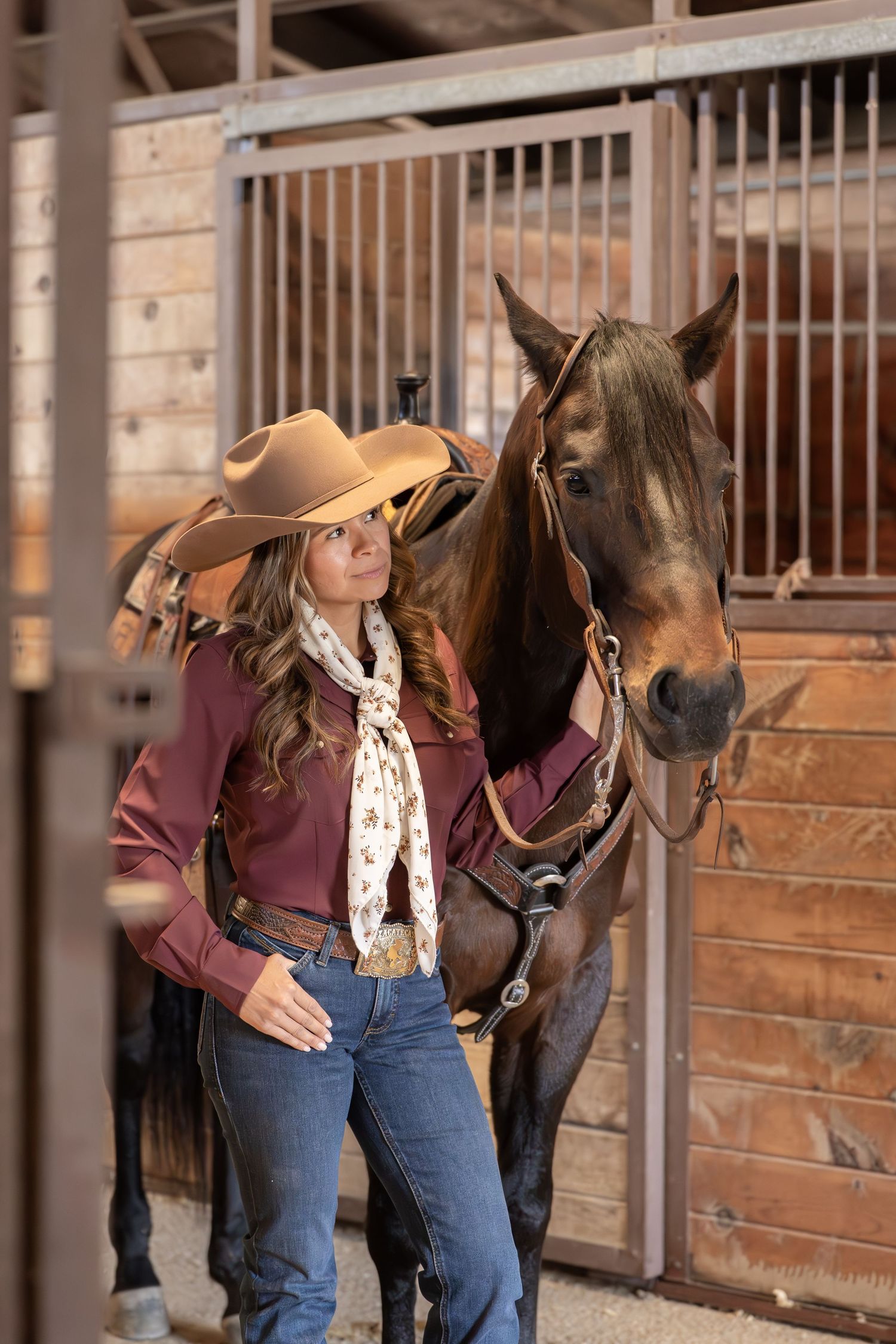 A cowgirl in a tan hat and burgundy jacket stands beside a saddled bay horse in a wooden stable.