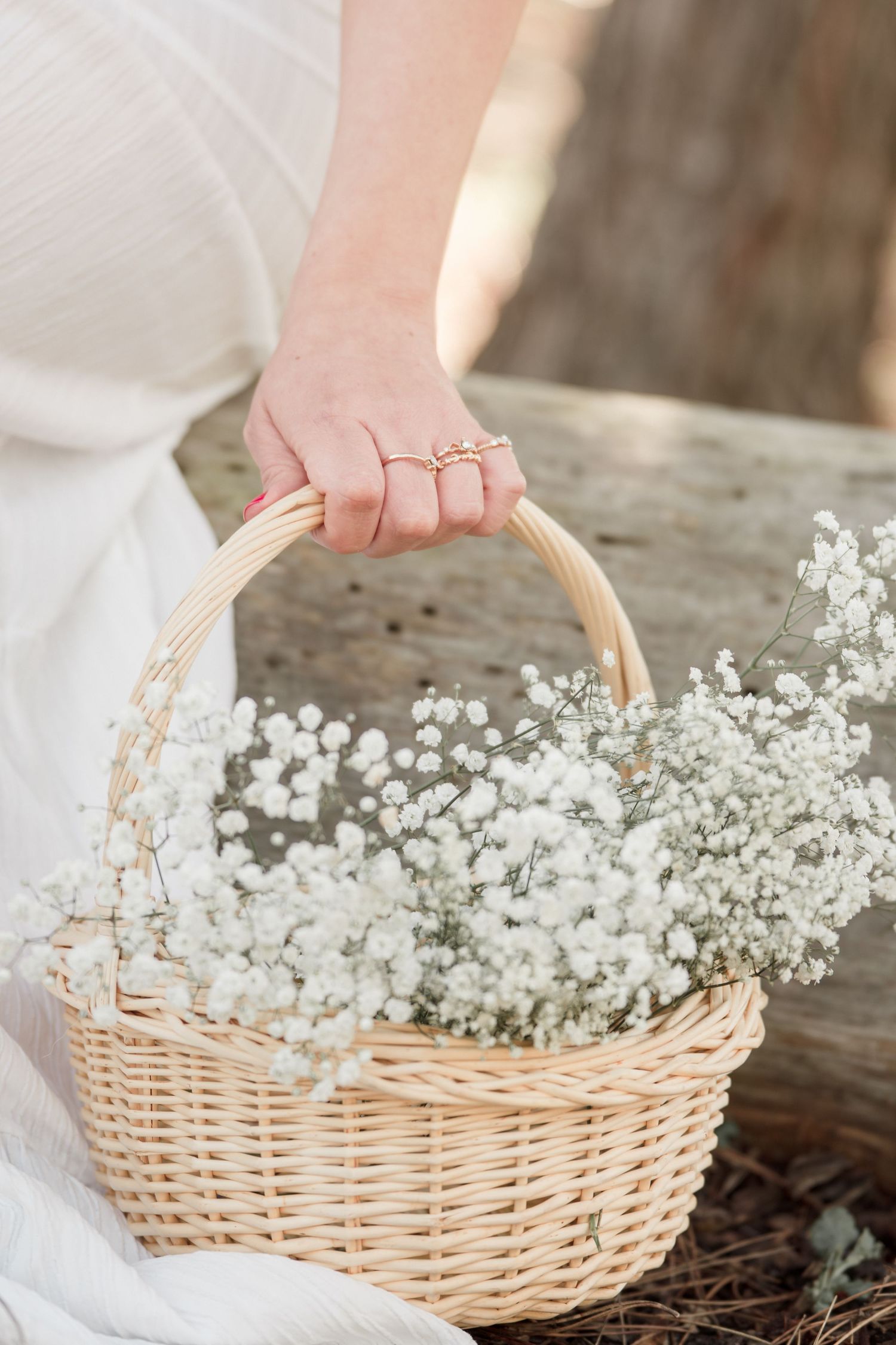 Woman in white dress holding a wicker basket filled with delicate white baby's breath flowers, wearing gold rings.