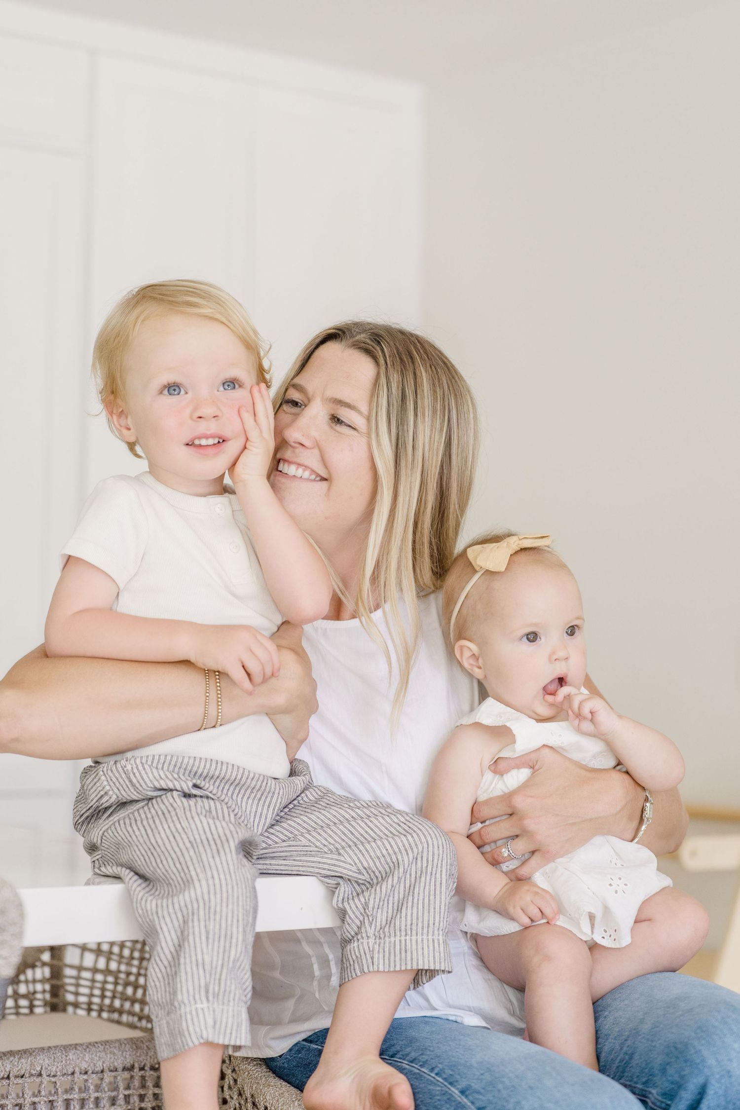 Smiling blonde mother sitting with toddler boy and baby girl in white outfits in a bright, airy room.