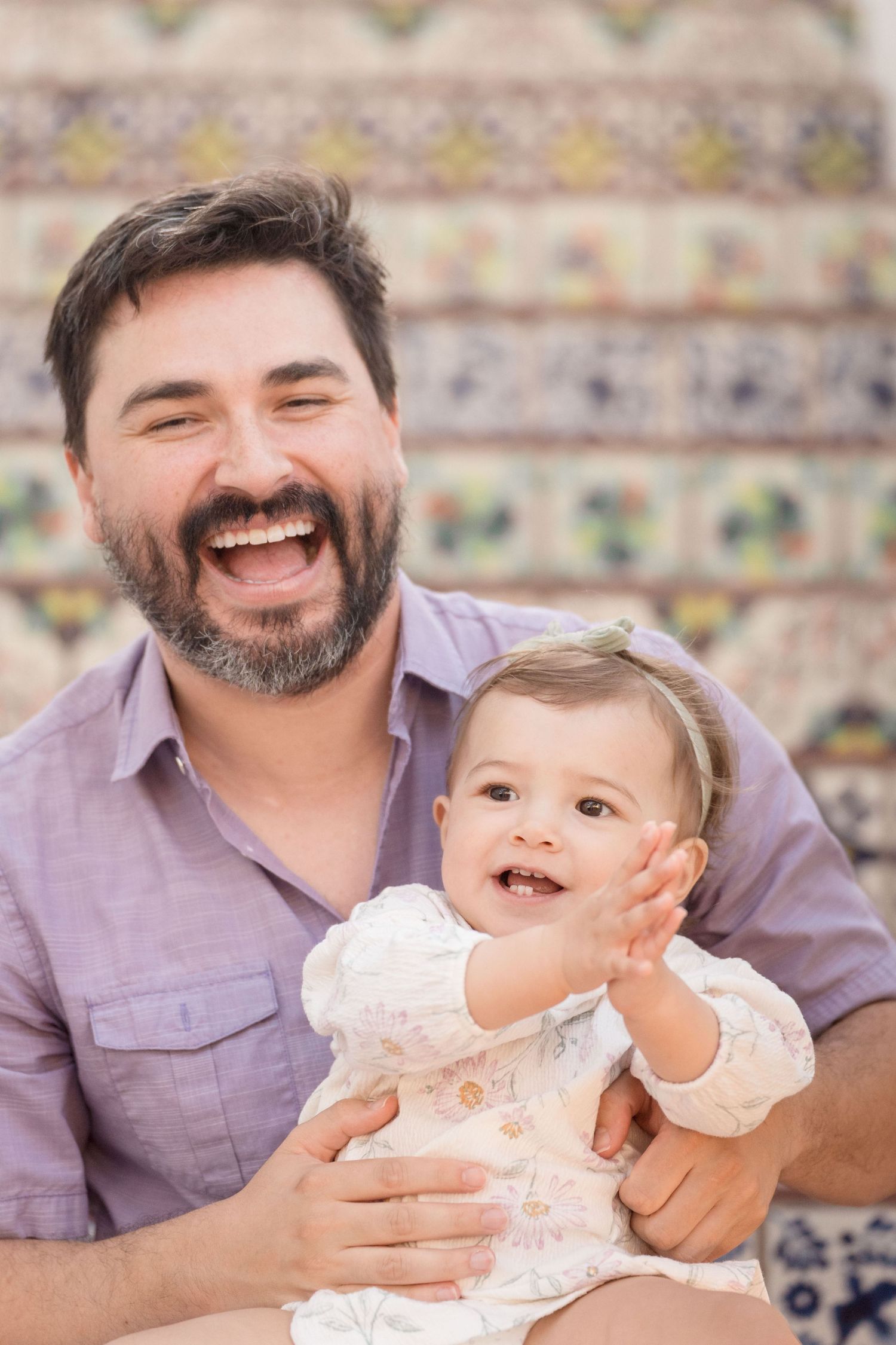 Laughing bearded father holding happy baby girl clapping hands, sitting in front of colorful tiled wall.