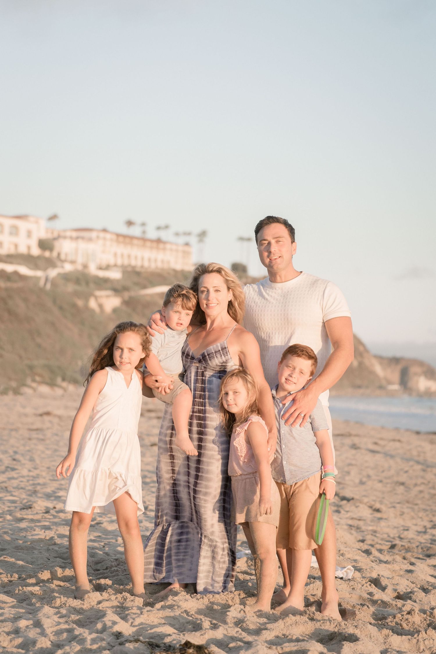 Happy family of six posing together on beach at sunset with coastal cliffs in background.