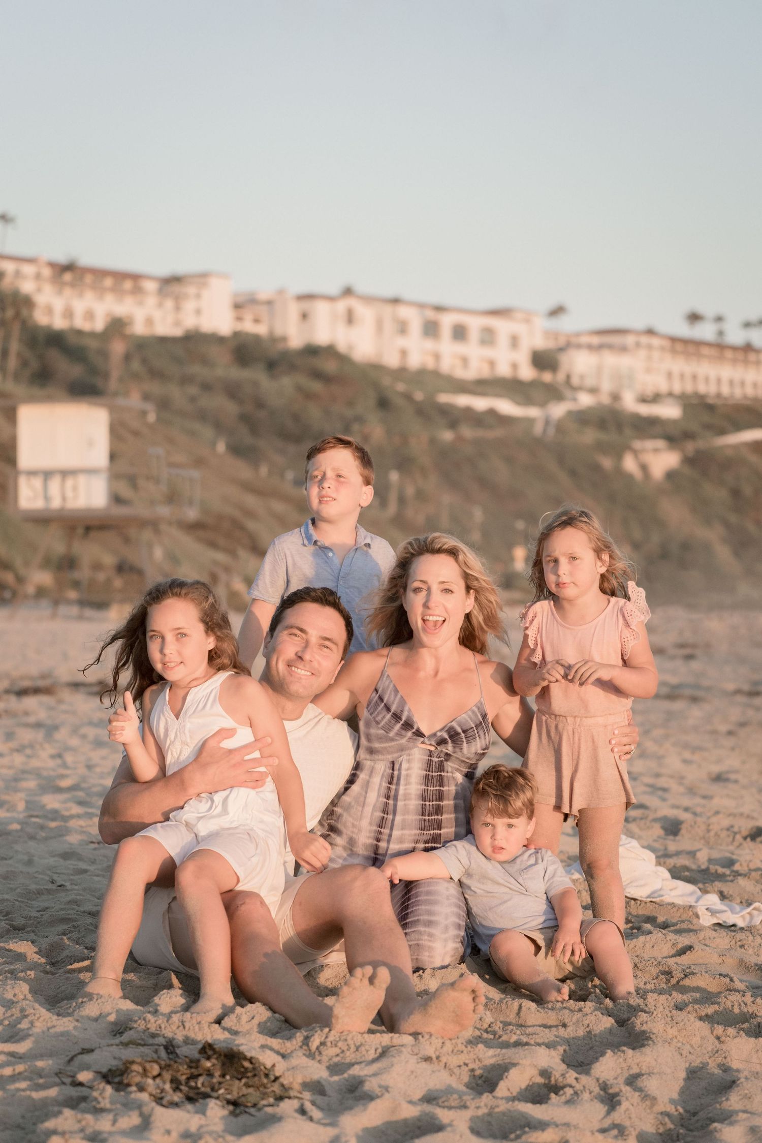 Smiling family of six sits together on a sandy beach with a cliffside resort in the background.