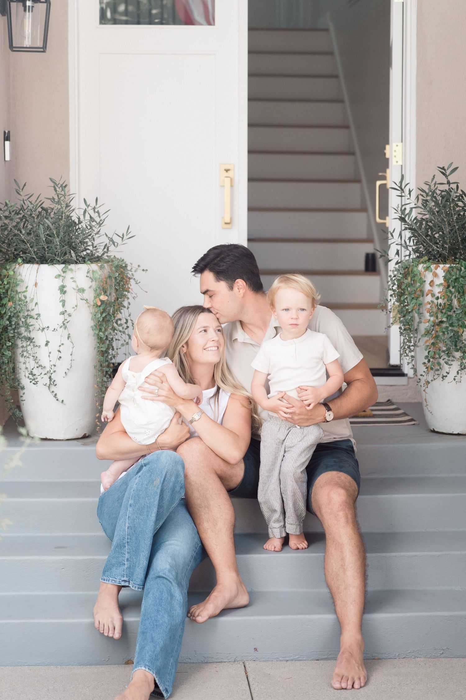 Young barefoot family of four sitting on front porch steps with two young children outdoors.