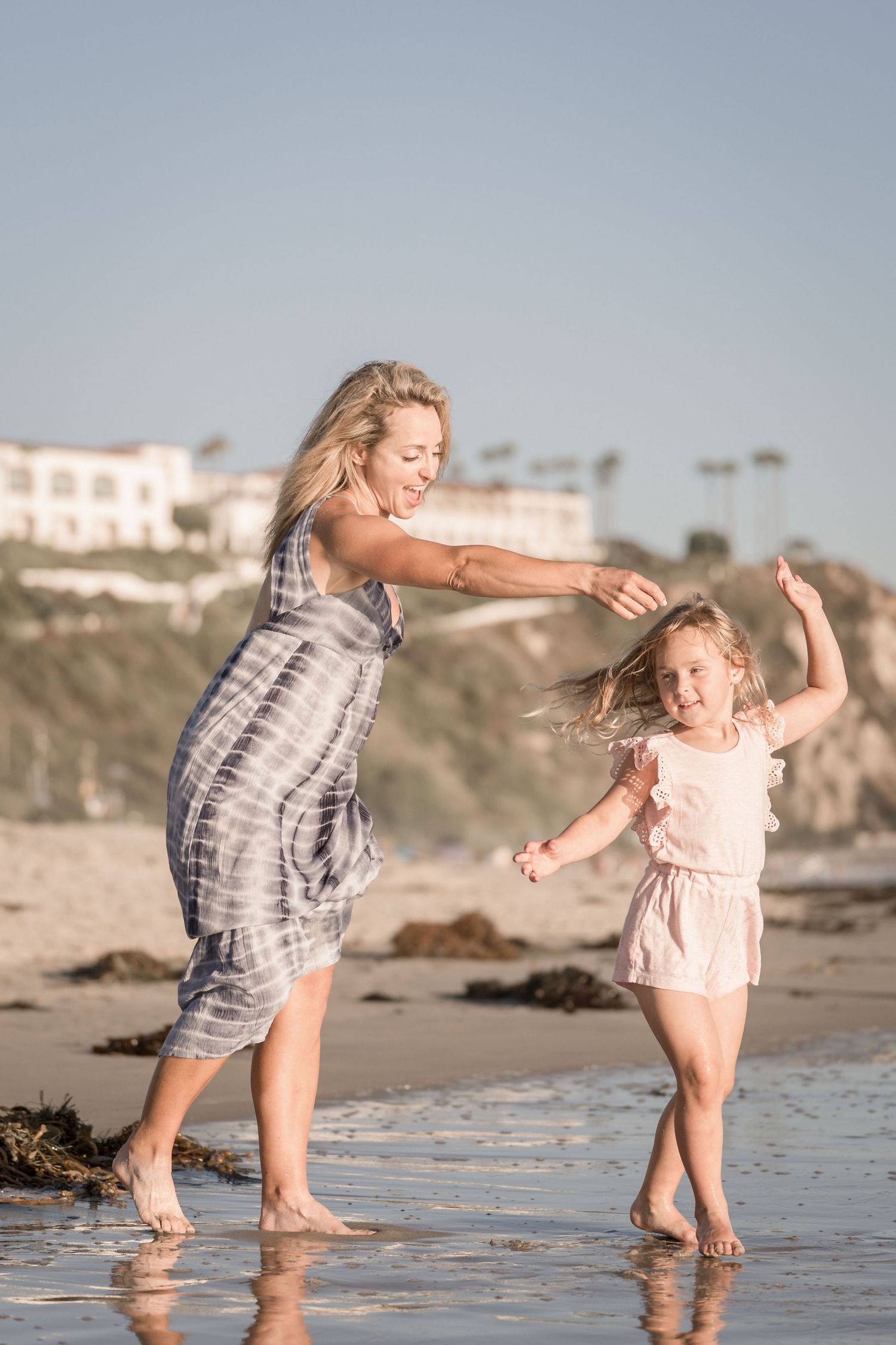 Mother and daughter dancing joyfully barefoot on a reflective beach at golden hour.