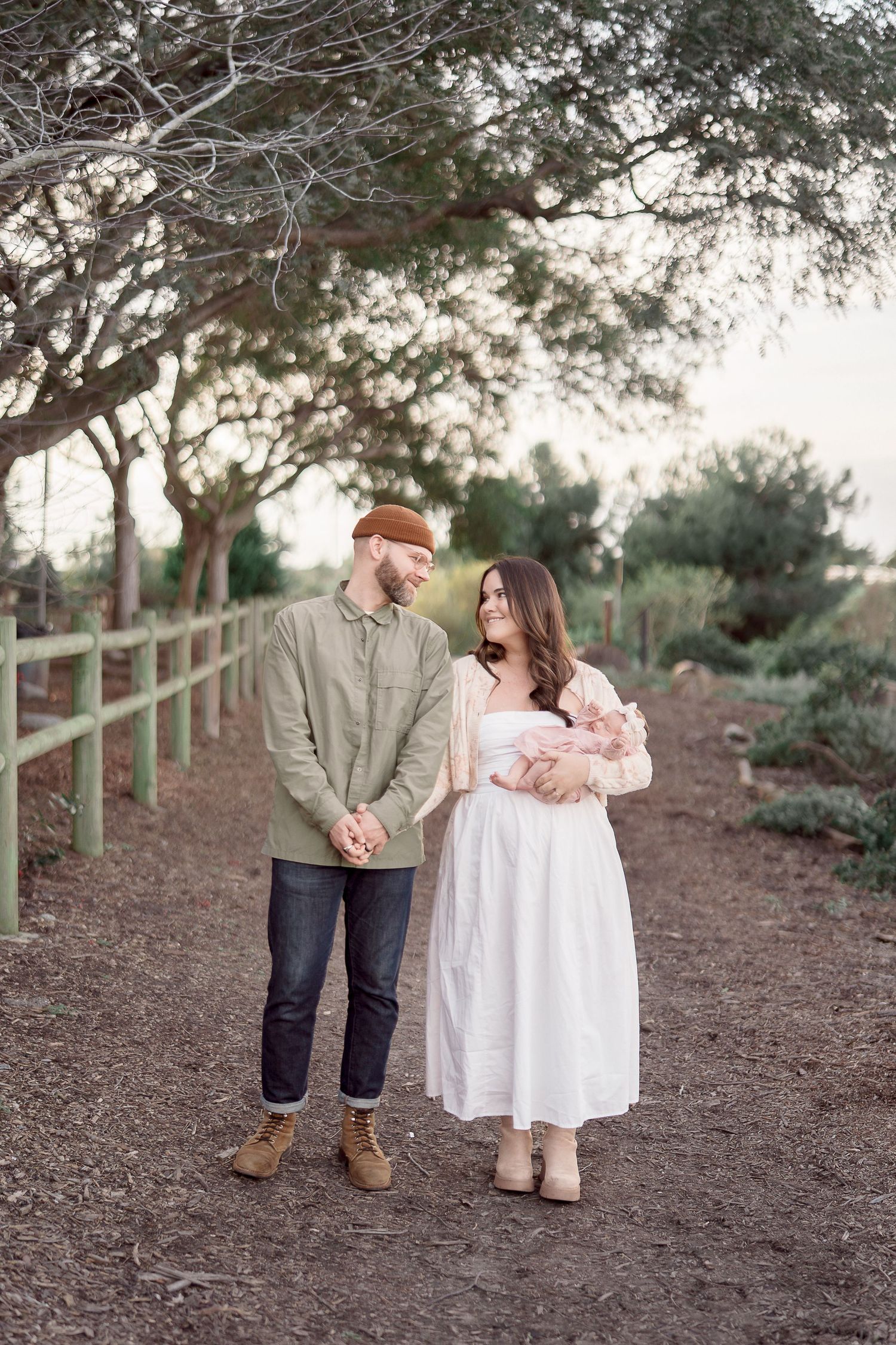 Young couple stands on a garden path holding their newborn baby, dressed in green and white outfits.