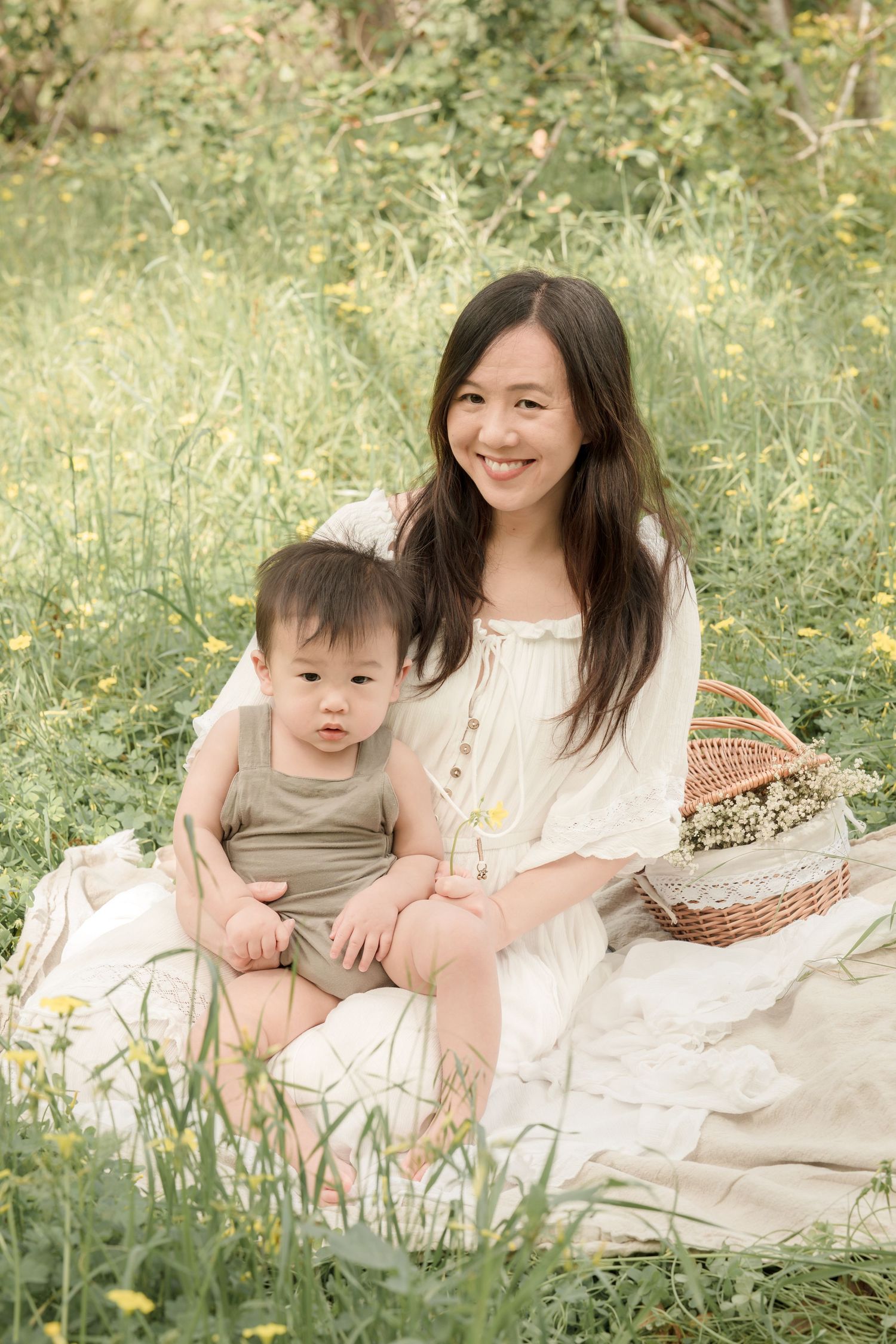 A cheerful Asian mother in a white dress sits on a blanket in a flower field with her baby and a wicker basket.