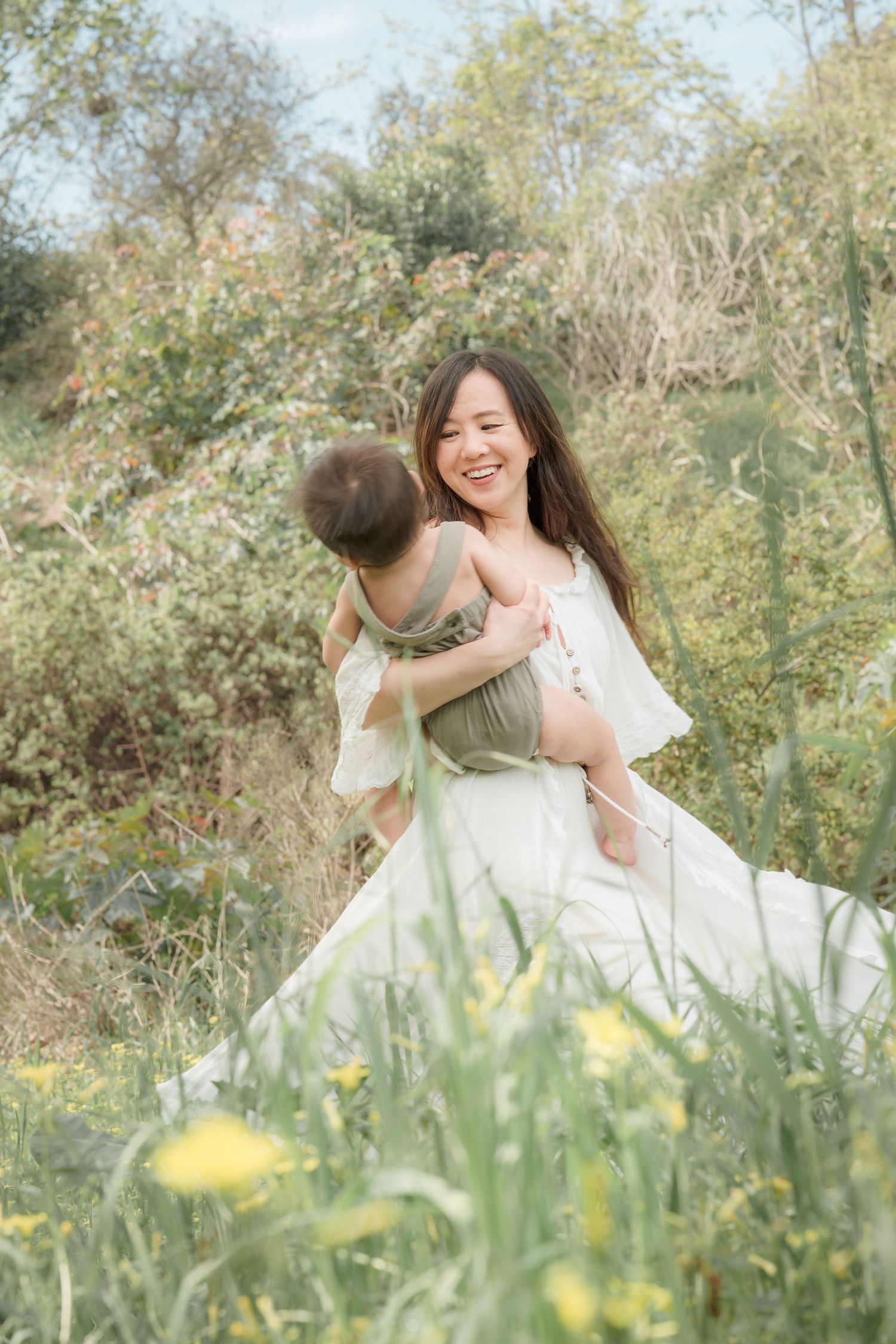 A joyful Asian mother in a white dress laughs while lifting her baby in a green romper in a sunny wildflower meadow.