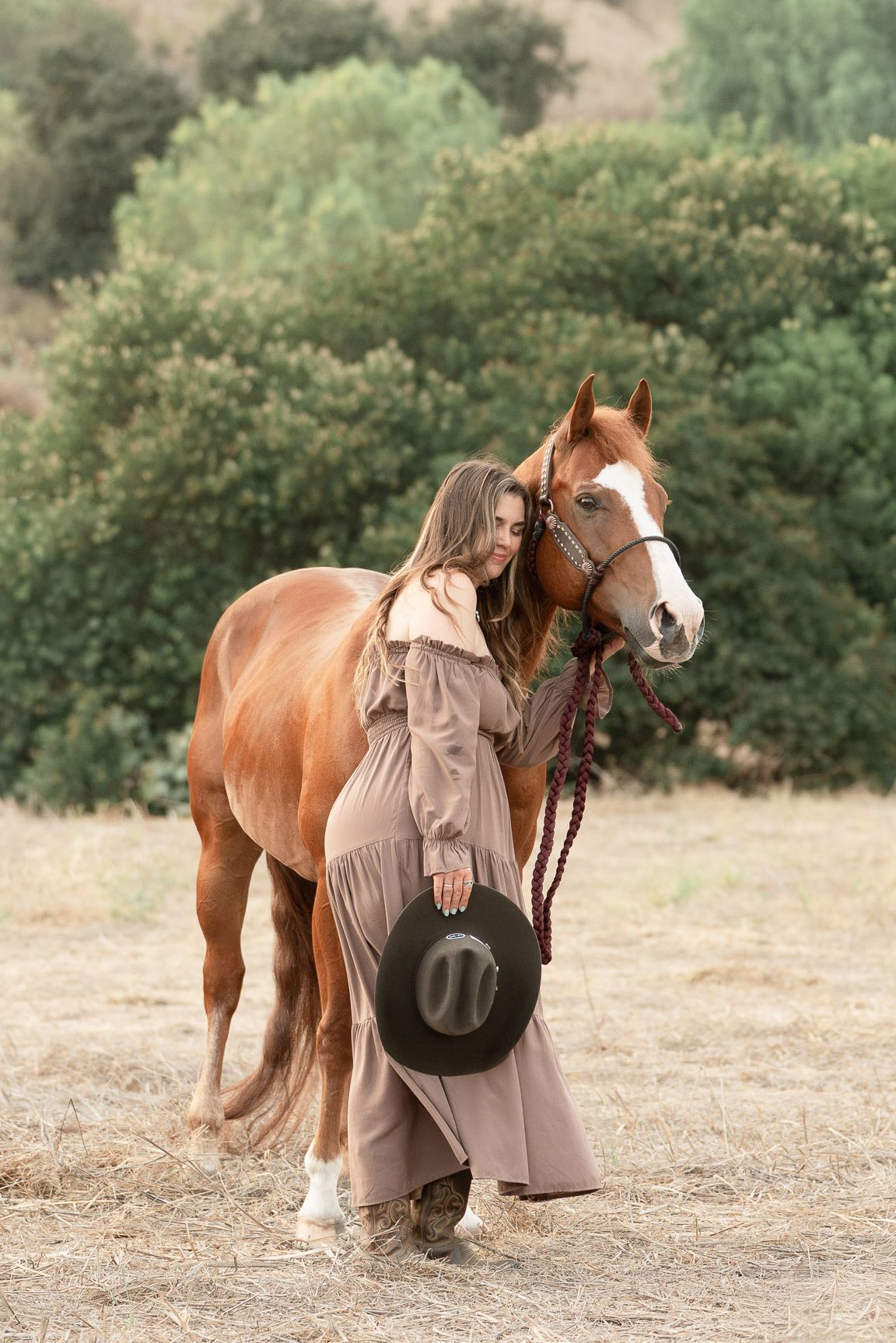 Woman in beige dress holding cowboy hat stands beside chestnut horse in a sunlit field with green trees.