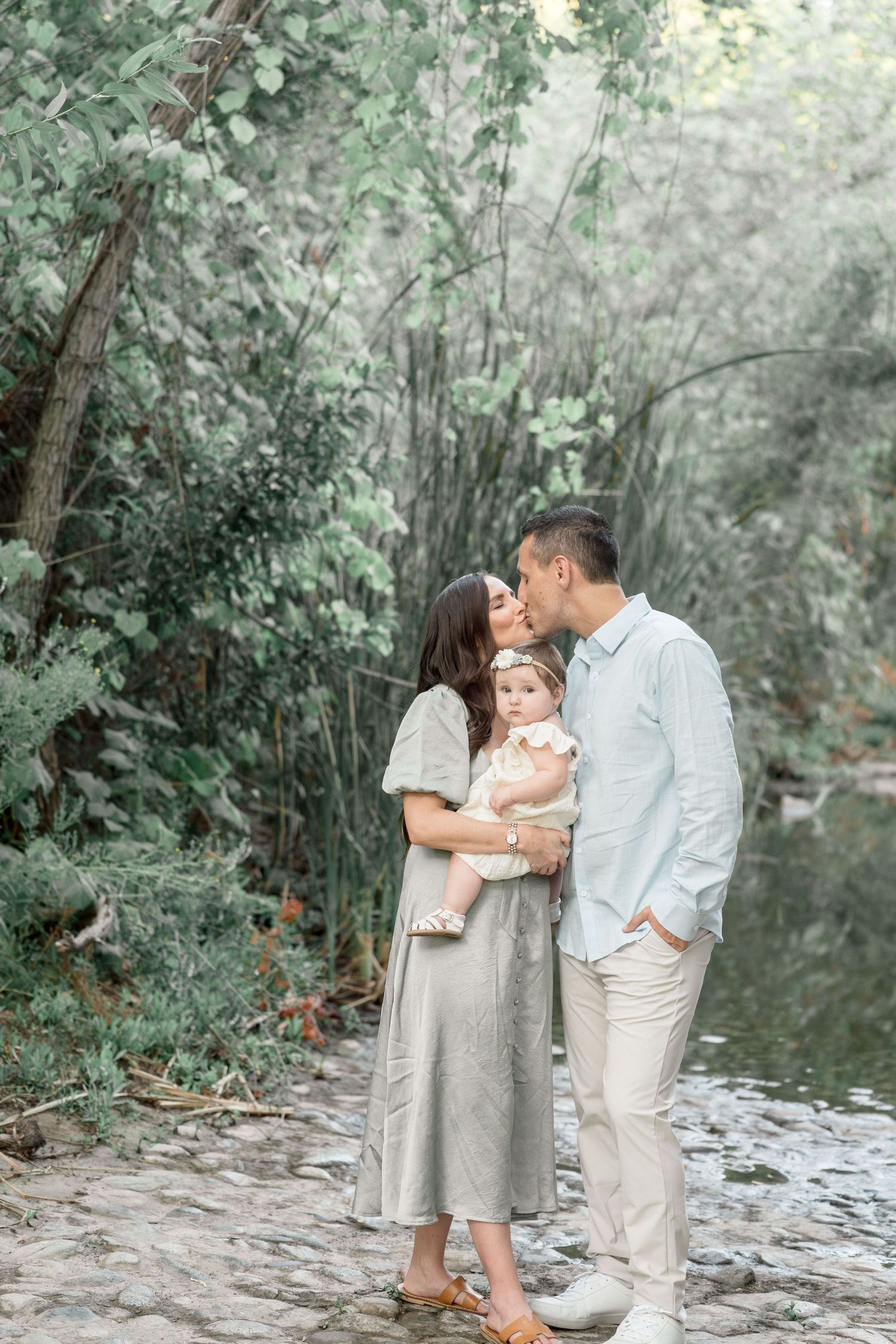 Family of three shares a kiss while holding baby girl near a lush green garden path.