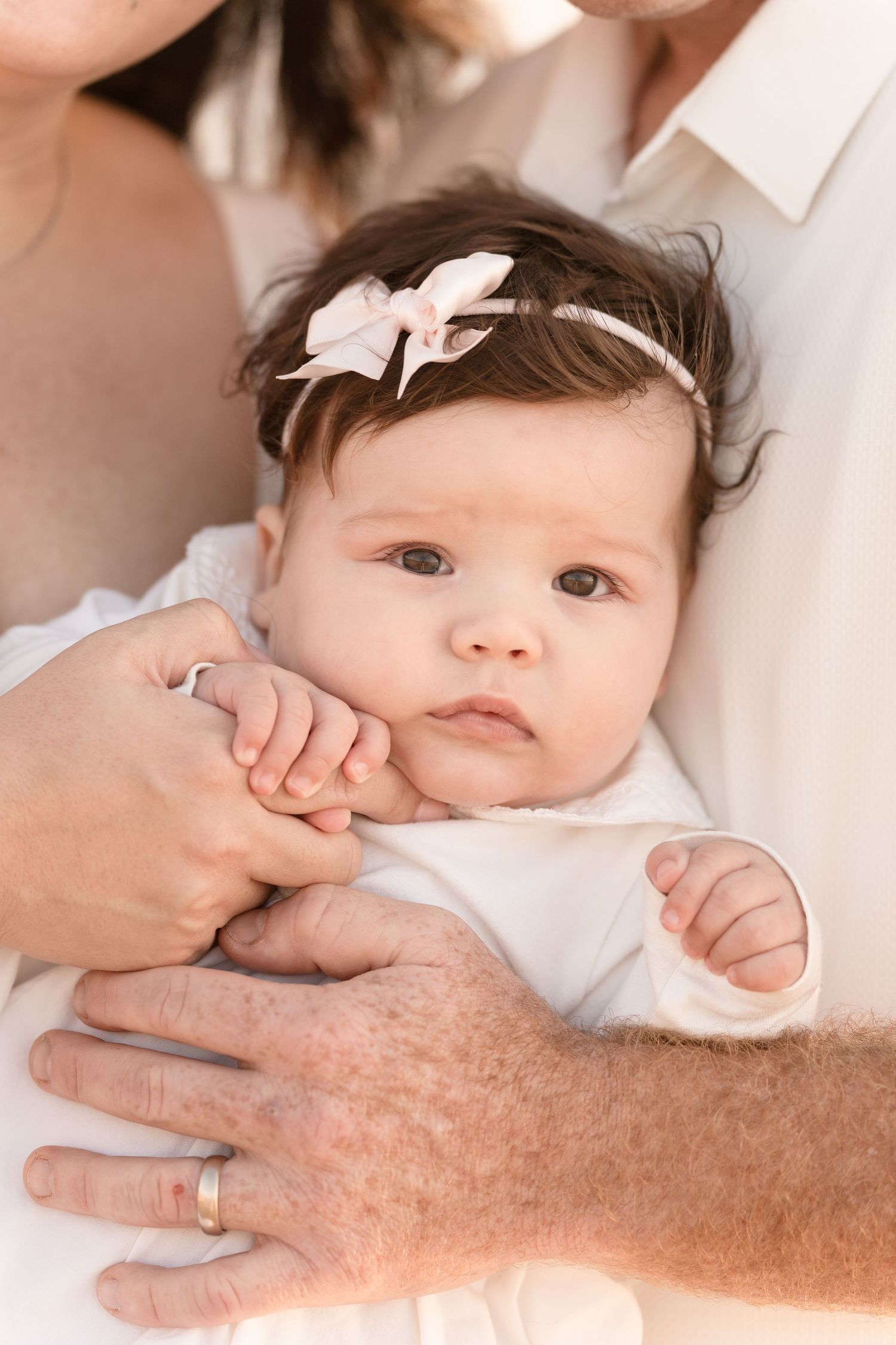 A newborn baby girl wearing a pink bow headband is cradled gently in her parents arms.