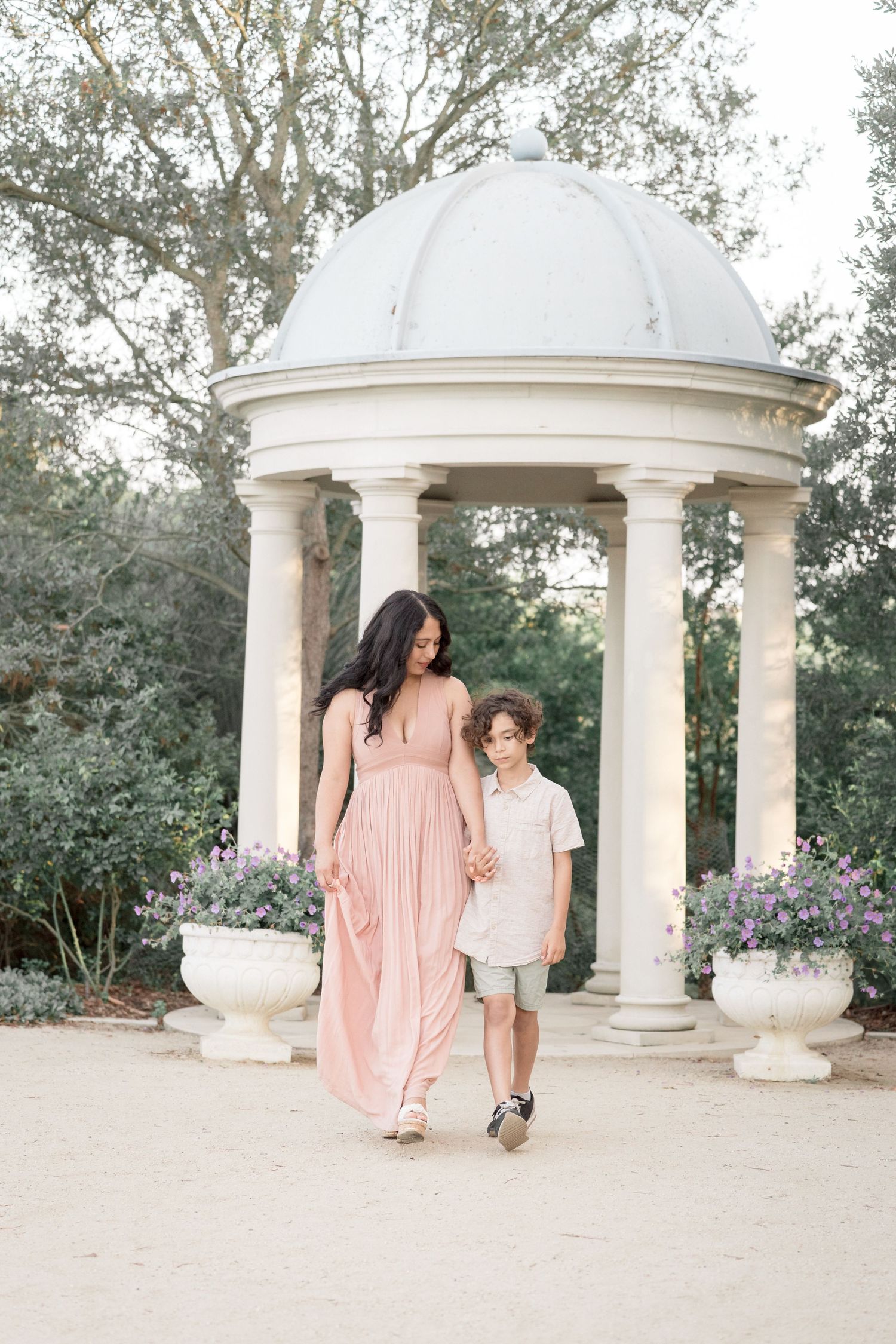 A mother in a blush maxi dress walks hand-in-hand with her young son near a white garden gazebo.