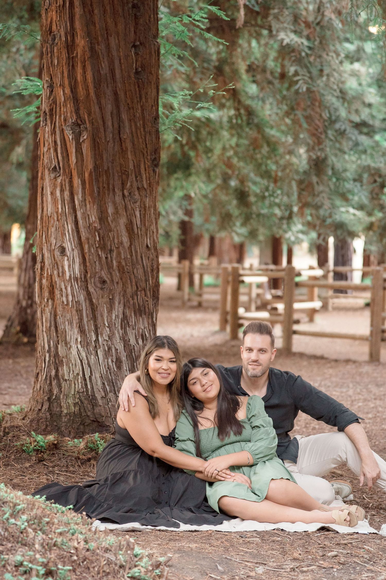Family of three sitting together against a large redwood tree in a serene forest park.
