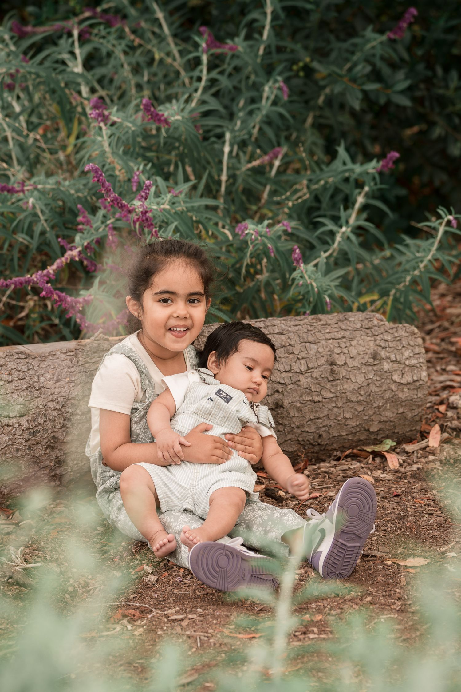 Two young siblings sit together on a log outdoors, surrounded by lush greenery and purple wildflowers.