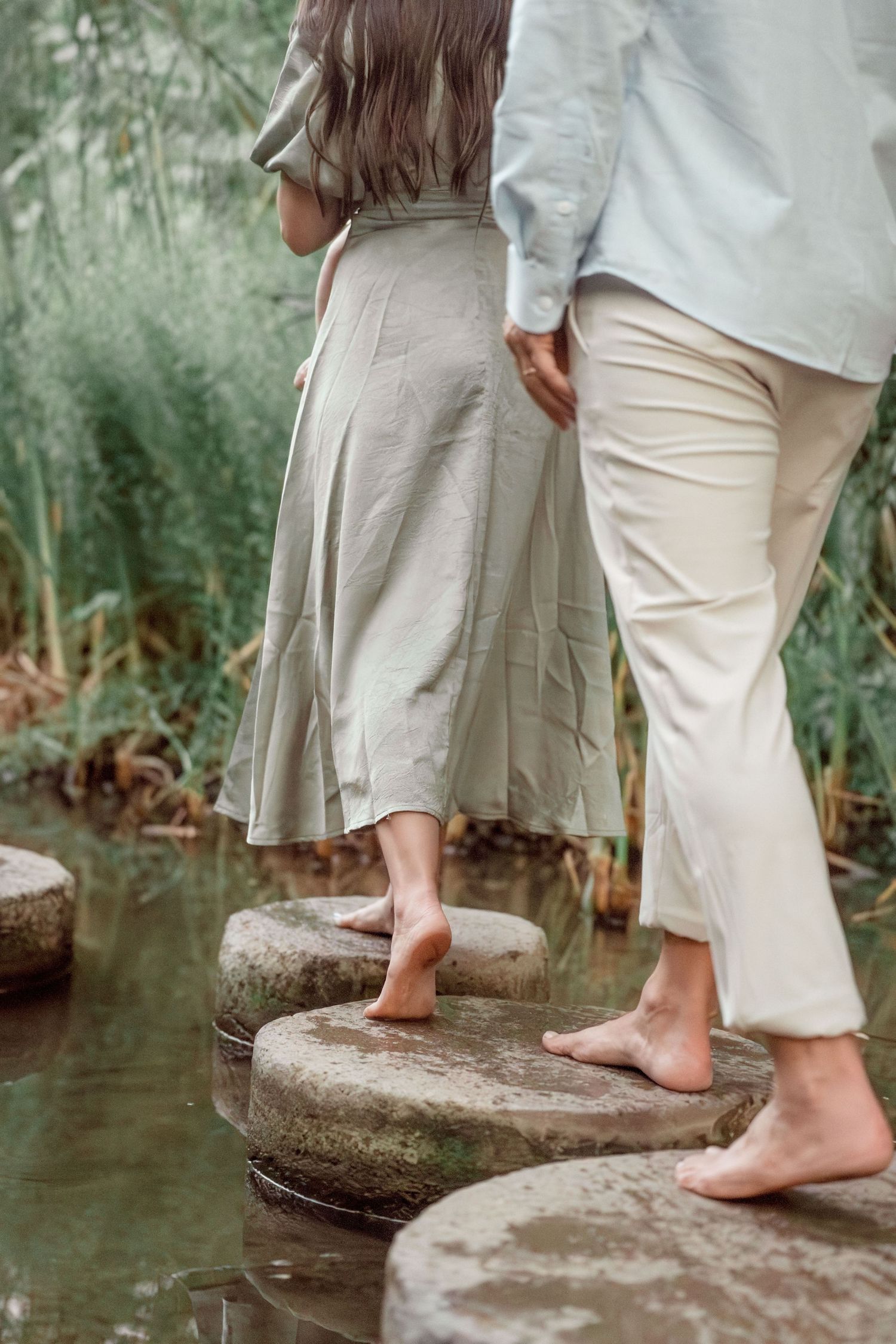 Barefoot couple in neutral tones walks on stepping stones over a peaceful stream in a lush green park.