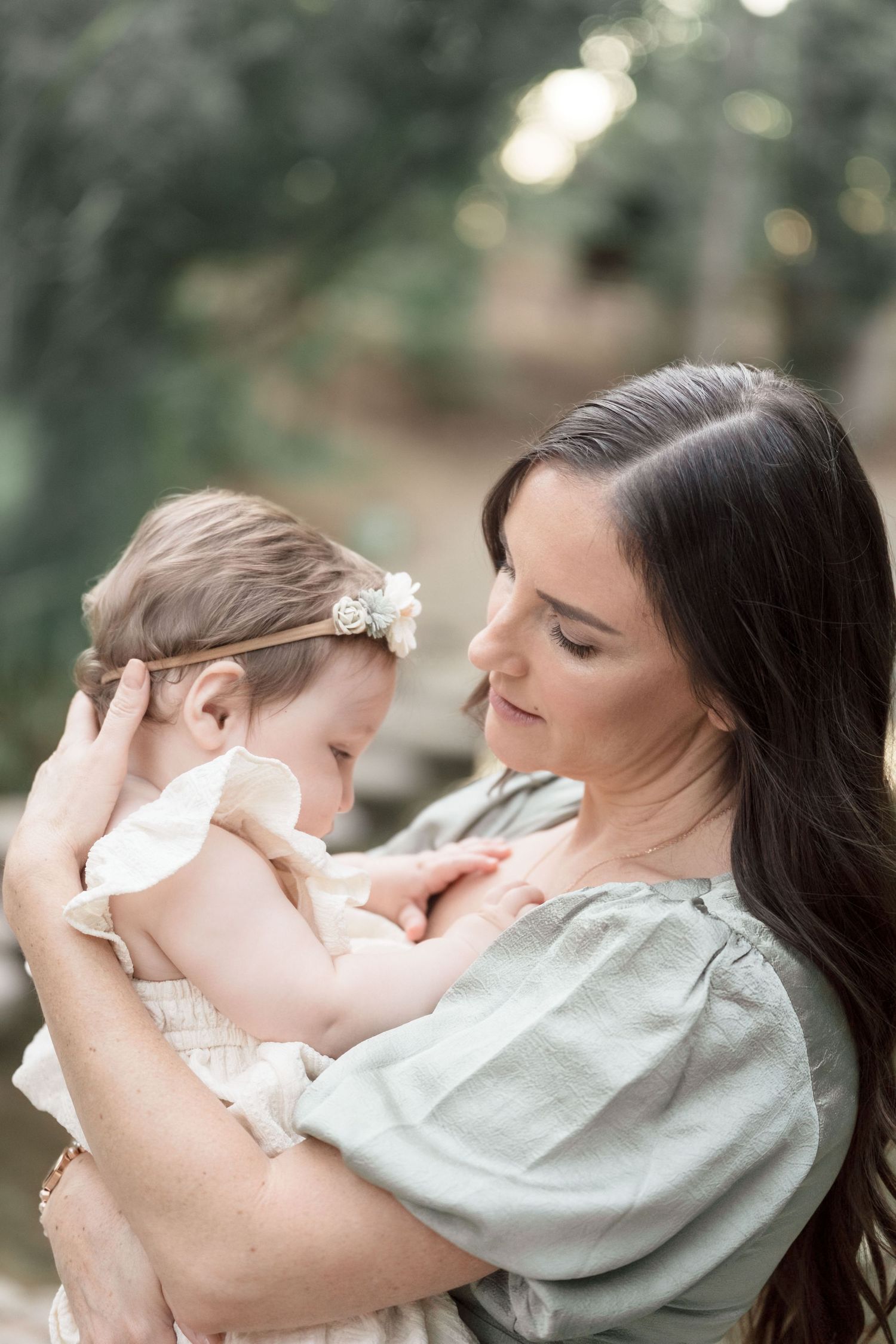 Tender moment as mother in sage dress gazes lovingly at baby girl with floral headband outdoors.